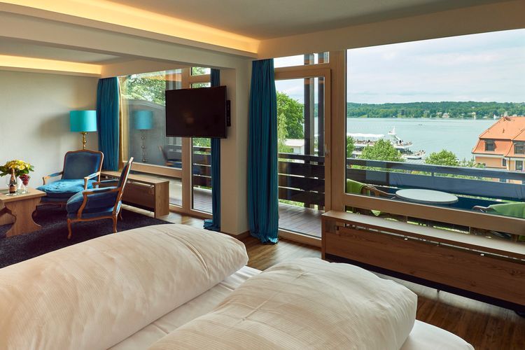 Hotel room with a view of a lake. White bedding, turquoise curtains, balcony, and a mounted TV.