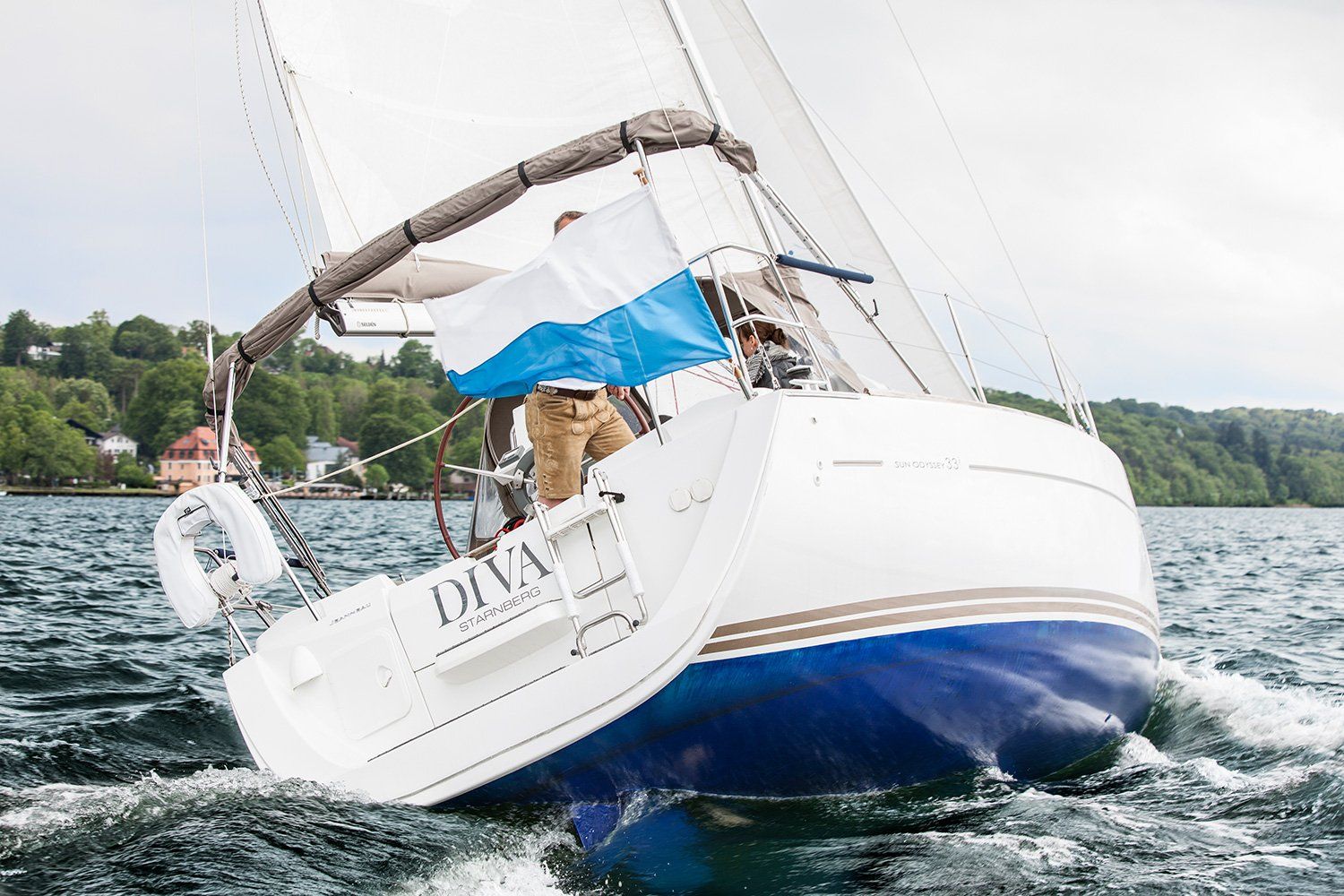 Das Segelboot „Diva“ liegt krängend im Wasser, am Steuer steht eine Person, und es weht eine blau-weiße Flagge.