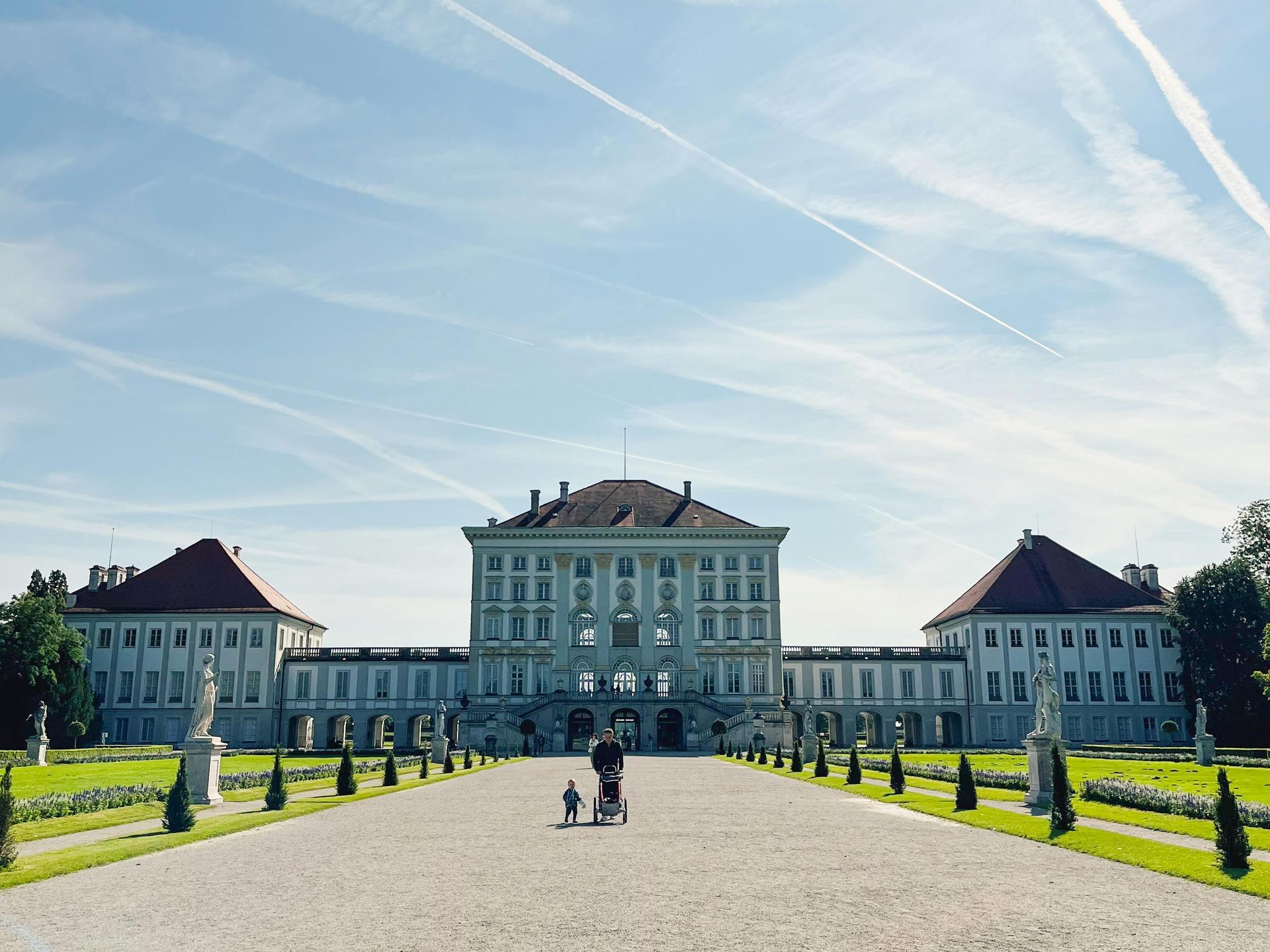 Ein Palast mit rotem Dach, flankiert von zwei Seitenflügeln; ein Kiesweg führt zum Eingang.