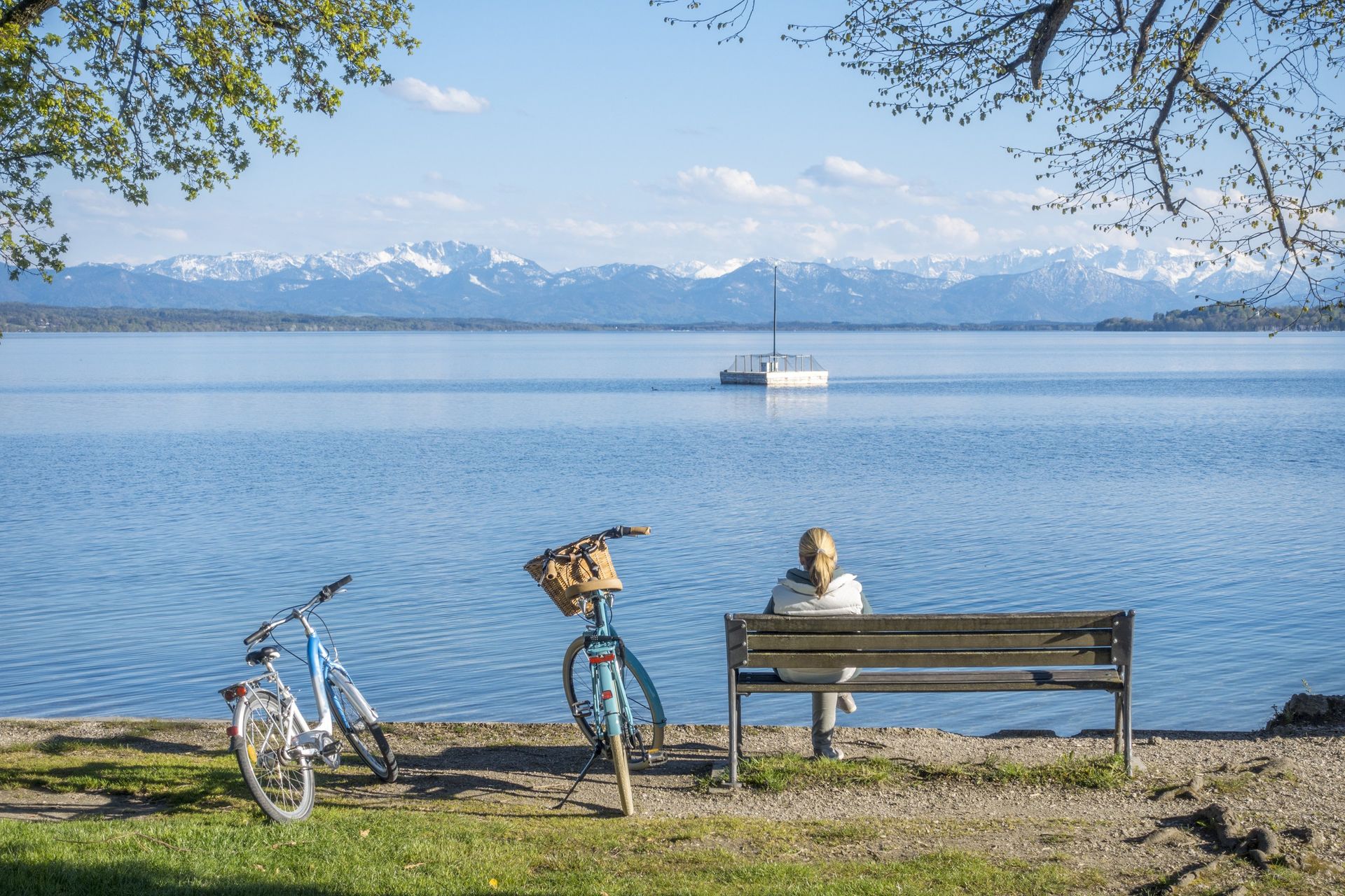 A person sits on a bench, looking at a calm lake and snow-capped mountains; bicycles rest beside the bench.