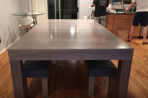 Modern dining table with benches in a kitchen; two people stand in the background.