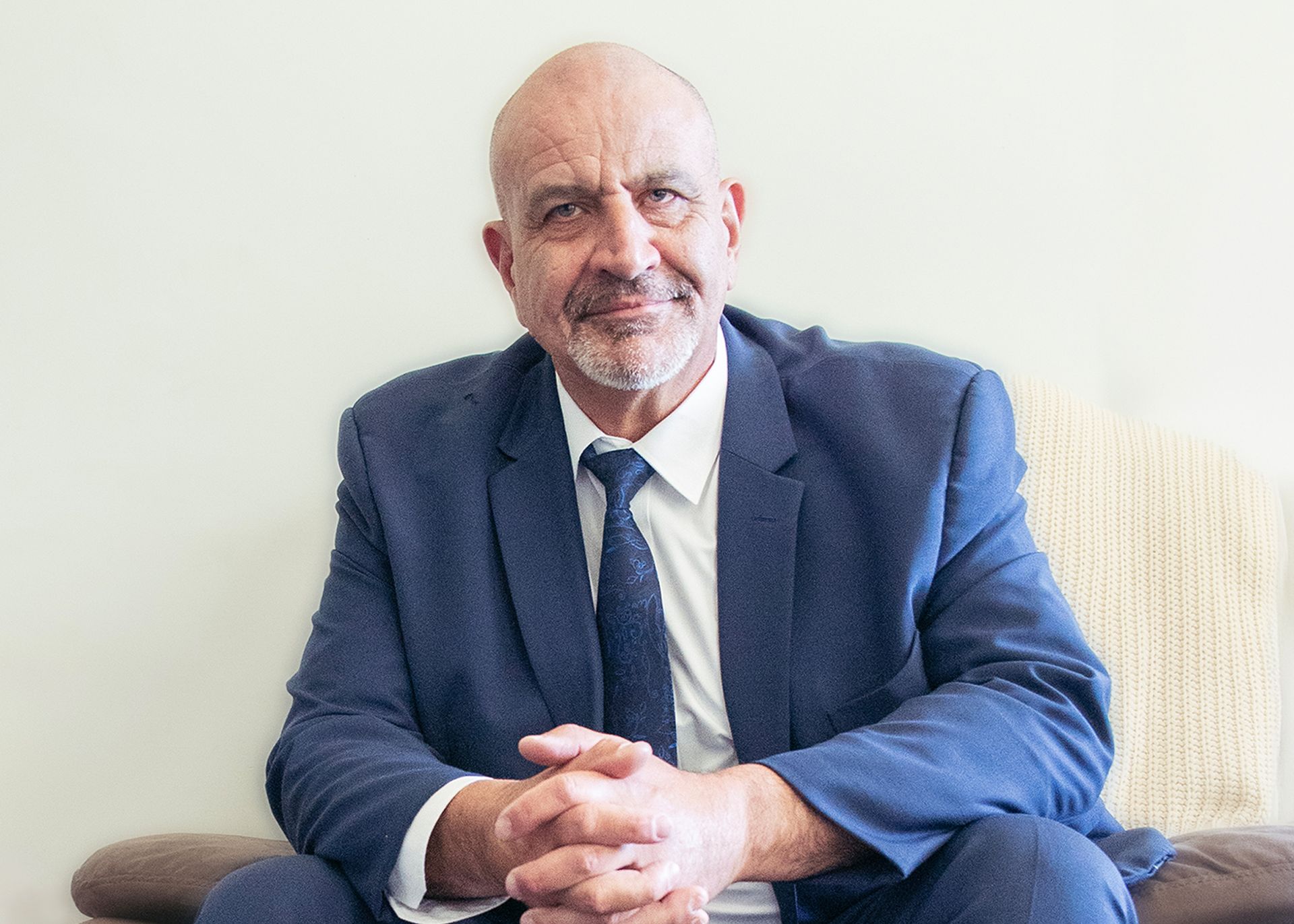 A man in a suit and tie is sitting on a couch with his hands folded.