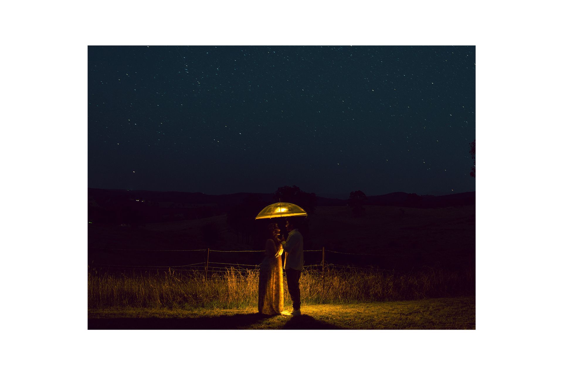 A bride and groom are standing under an umbrella in a field at night.
