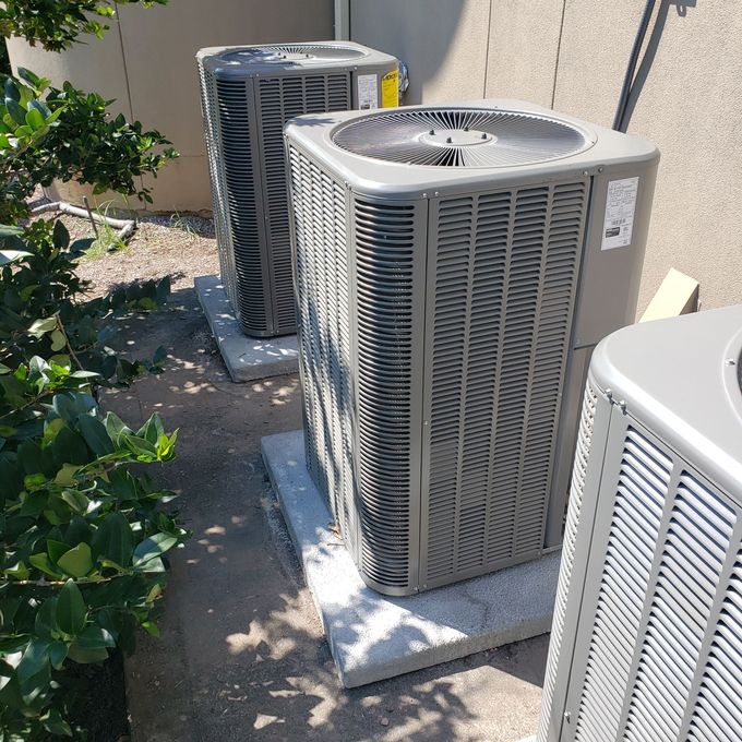 Three outdoor HVAC units beside a wall, with one in the foreground on a concrete pad.