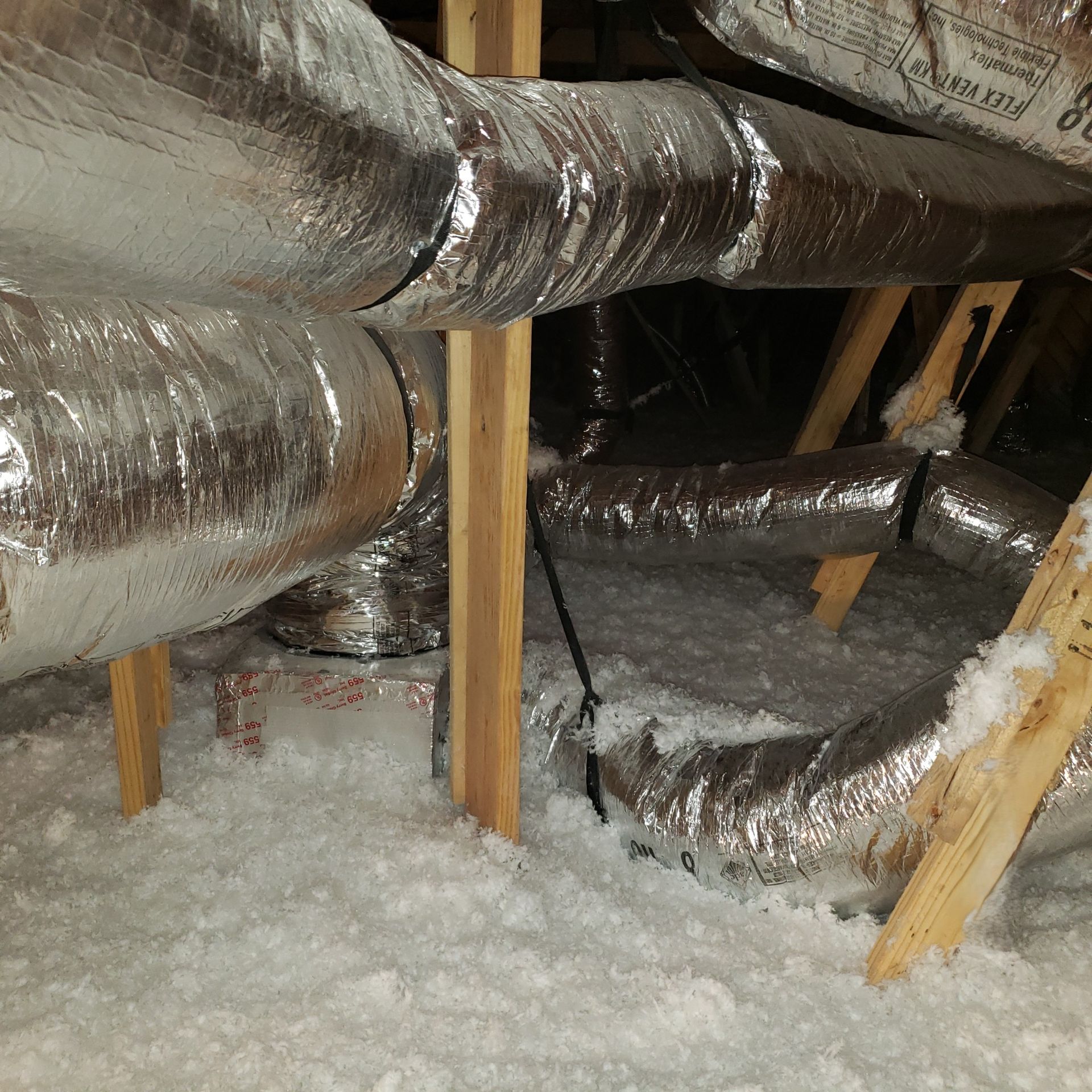 Insulated HVAC ducts in an attic with exposed wooden supports and loose white insulation.