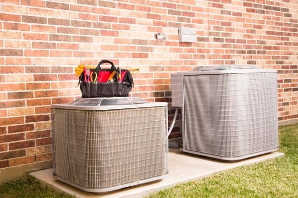 Two outdoor air conditioning units beside a brick wall, with a tool bag on the left unit.