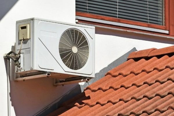 Outdoor air conditioner unit mounted on a wall beside a red tile roof and window blinds