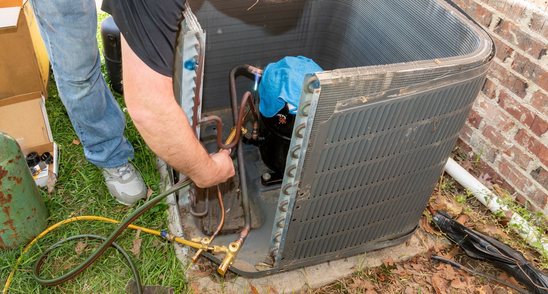 Technician repairing an outdoor HVAC unit beside a brick wall, with hoses and tools visible.