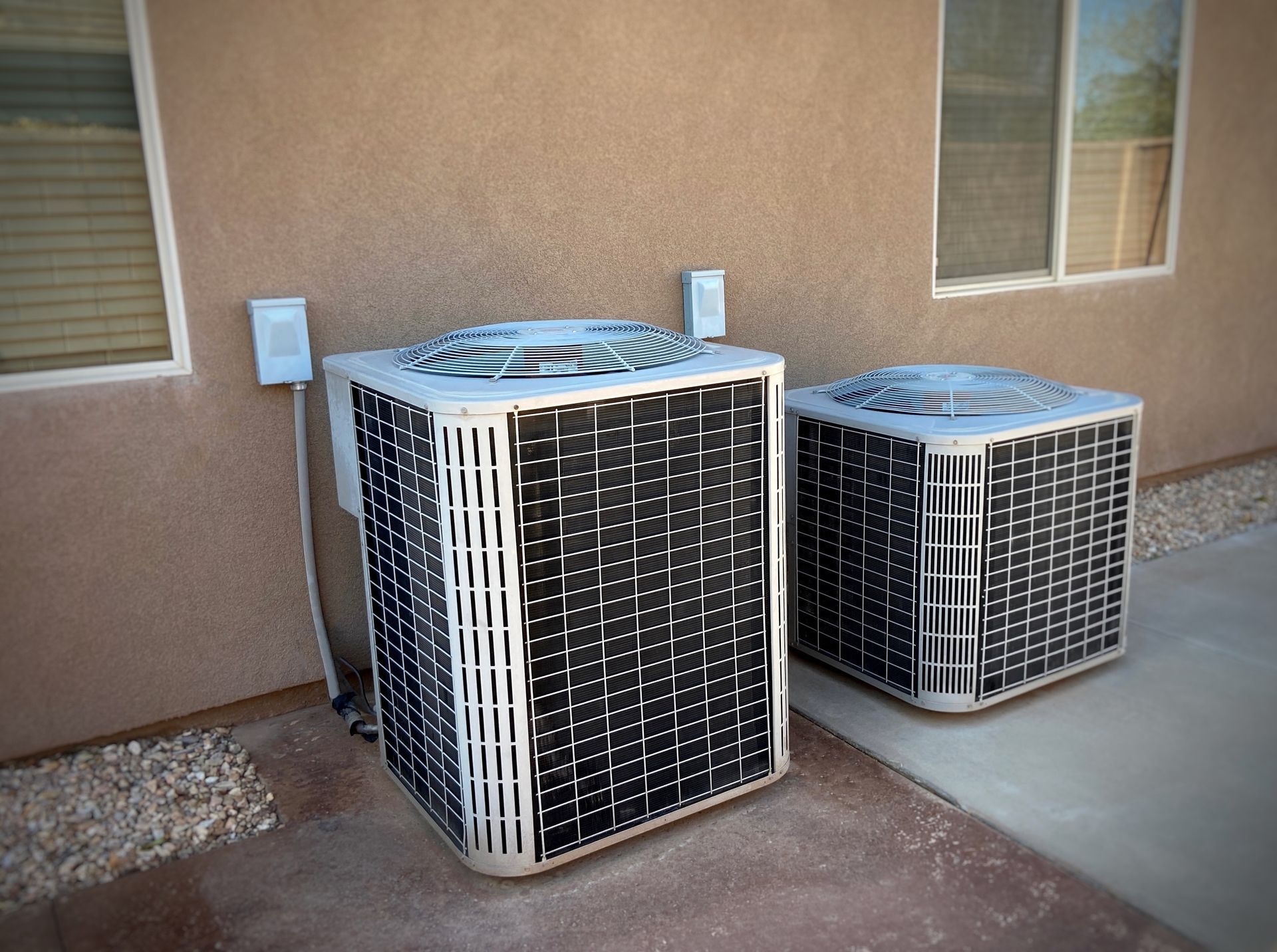 Two outdoor air conditioning units beside a beige house wall on a concrete patio.
