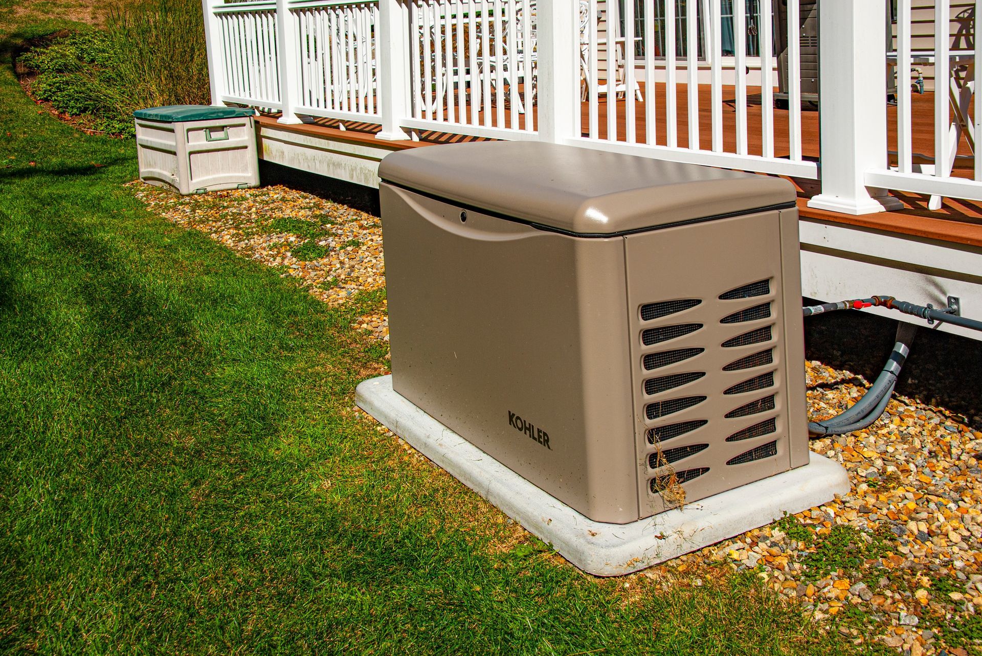 Outdoor beige generator beside a white deck railing on a gravel pad, with grass nearby