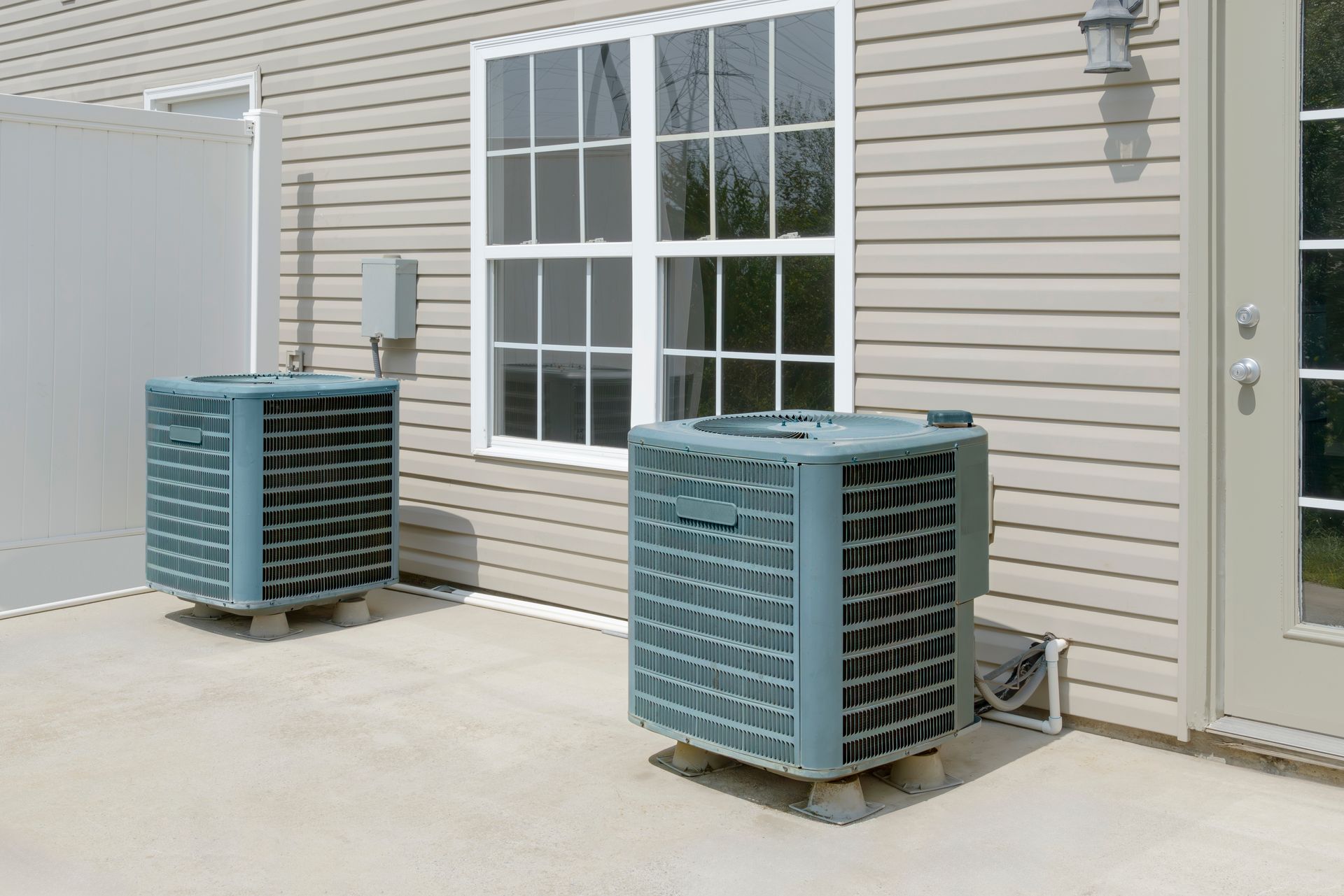Two outdoor HVAC units beside a beige house wall and window on a concrete patio