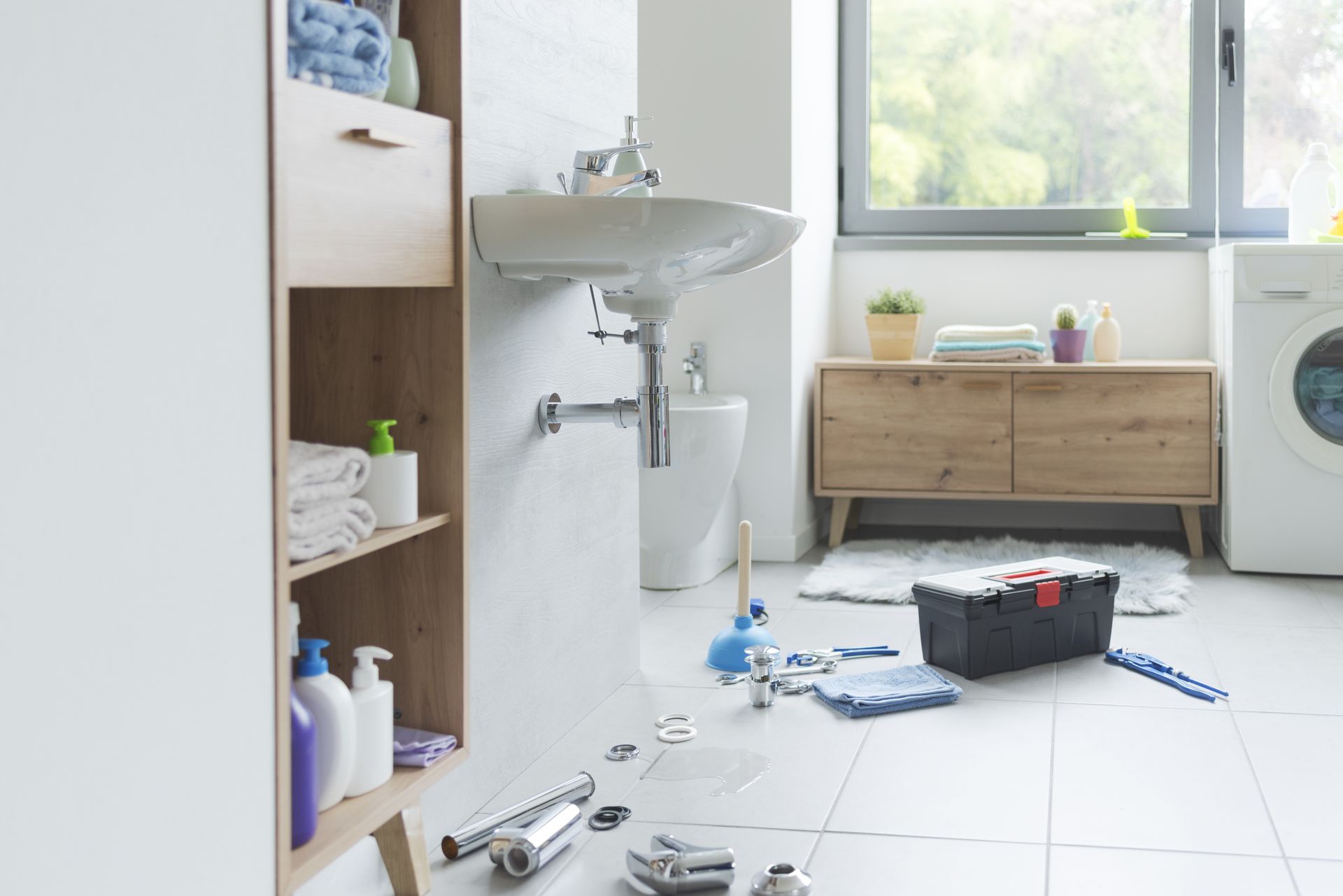 Cluttered bathroom floor with tools, cleaning supplies, and a black toolbox near a sink and window