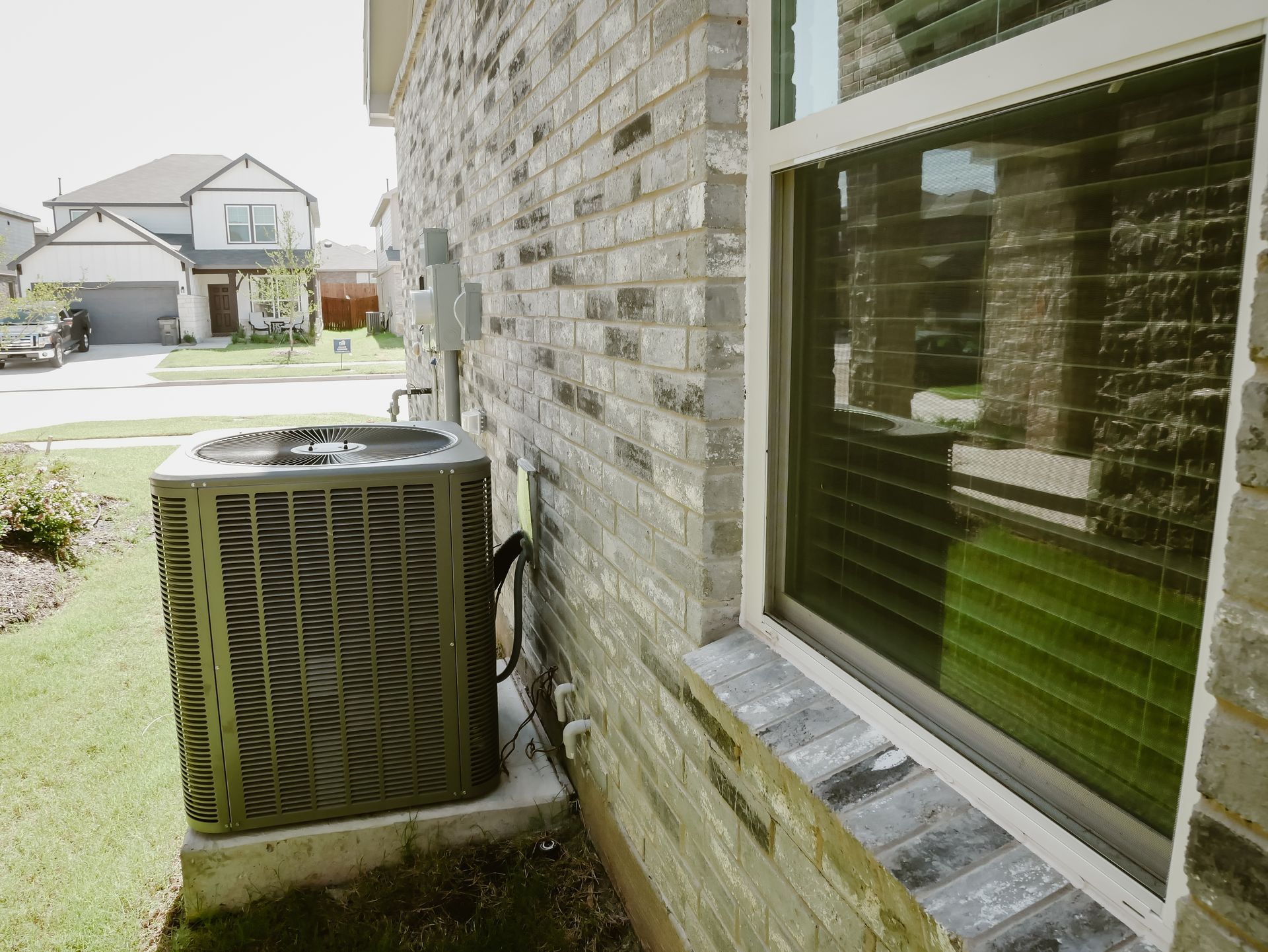 Side of a brick house with a window and air conditioner unit beside a lawn and street.