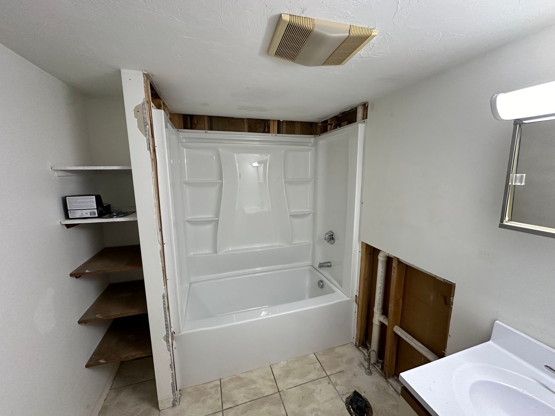 Bathroom renovation in progress, featuring a new white tub/shower unit. Exposed wall studs, shelving, and partially tiled floor are visible.