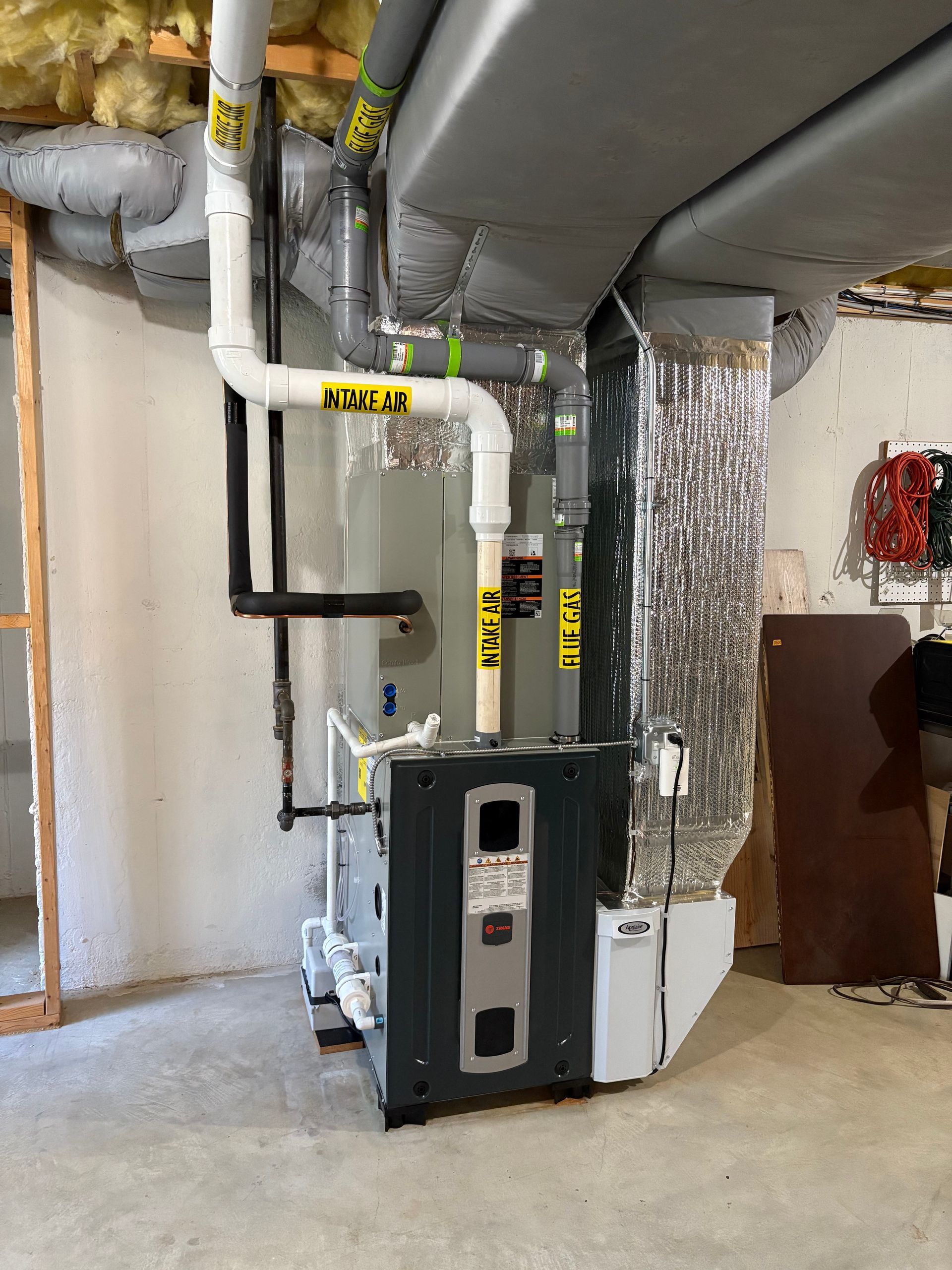 Furnace unit with ductwork in a basement setting. Pipes are white and black. Ductwork is silver.