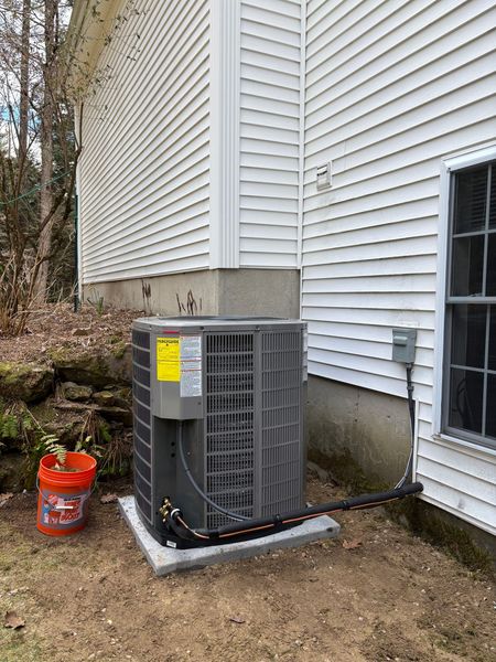Exterior view of an air conditioning unit next to a white house with a window. Orange bucket nearby.