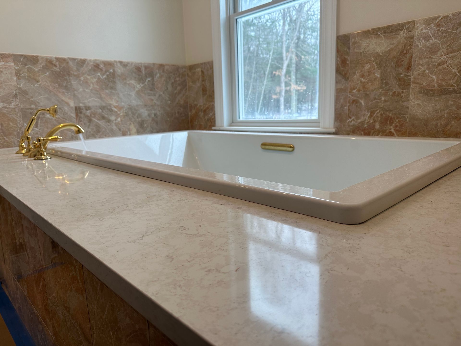 Bathtub set in countertop, gold faucet, marble tile backsplash, window.