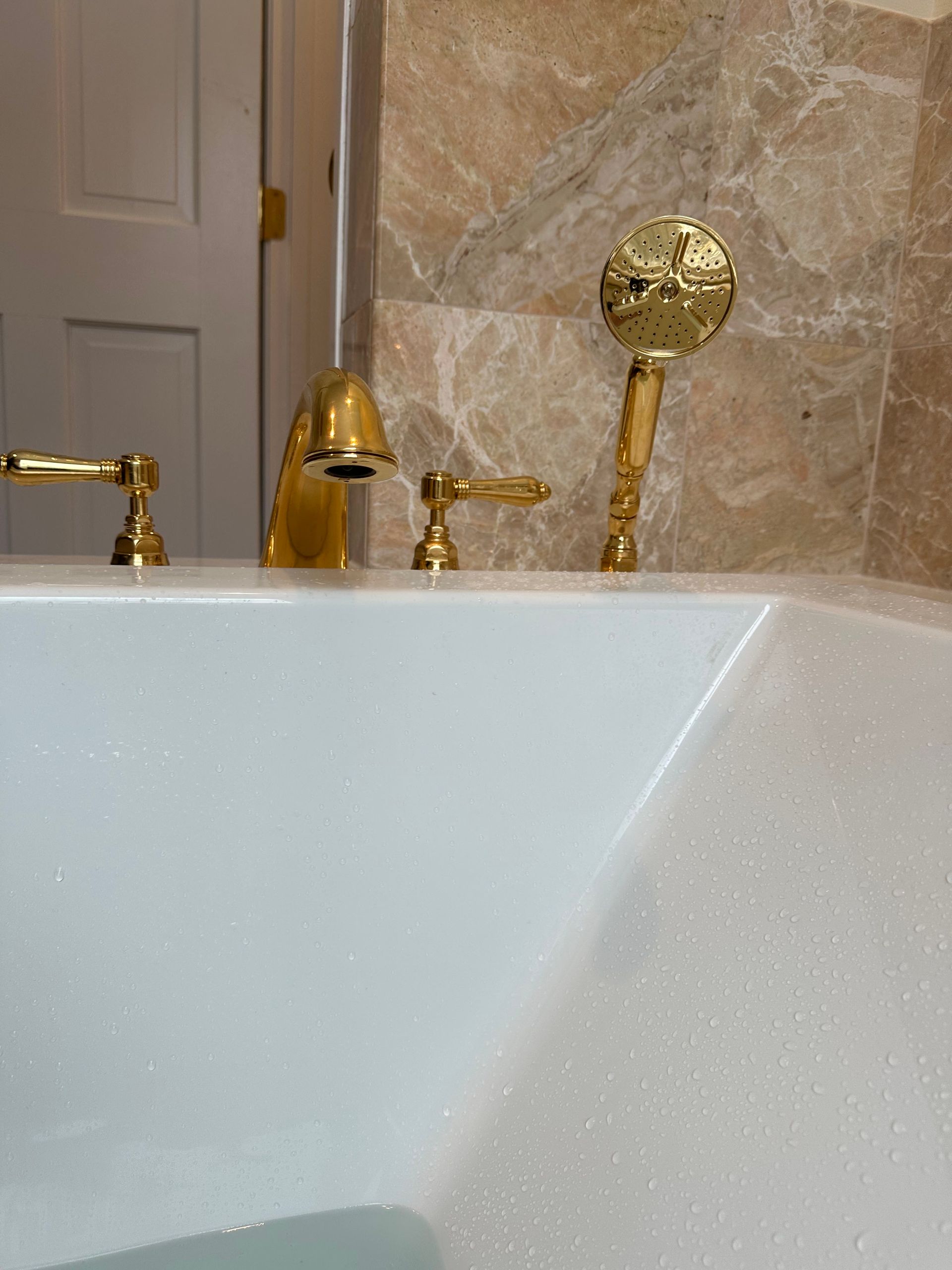 Gold fixtures on a white bathtub with water flowing. Beige marble wall in the background.