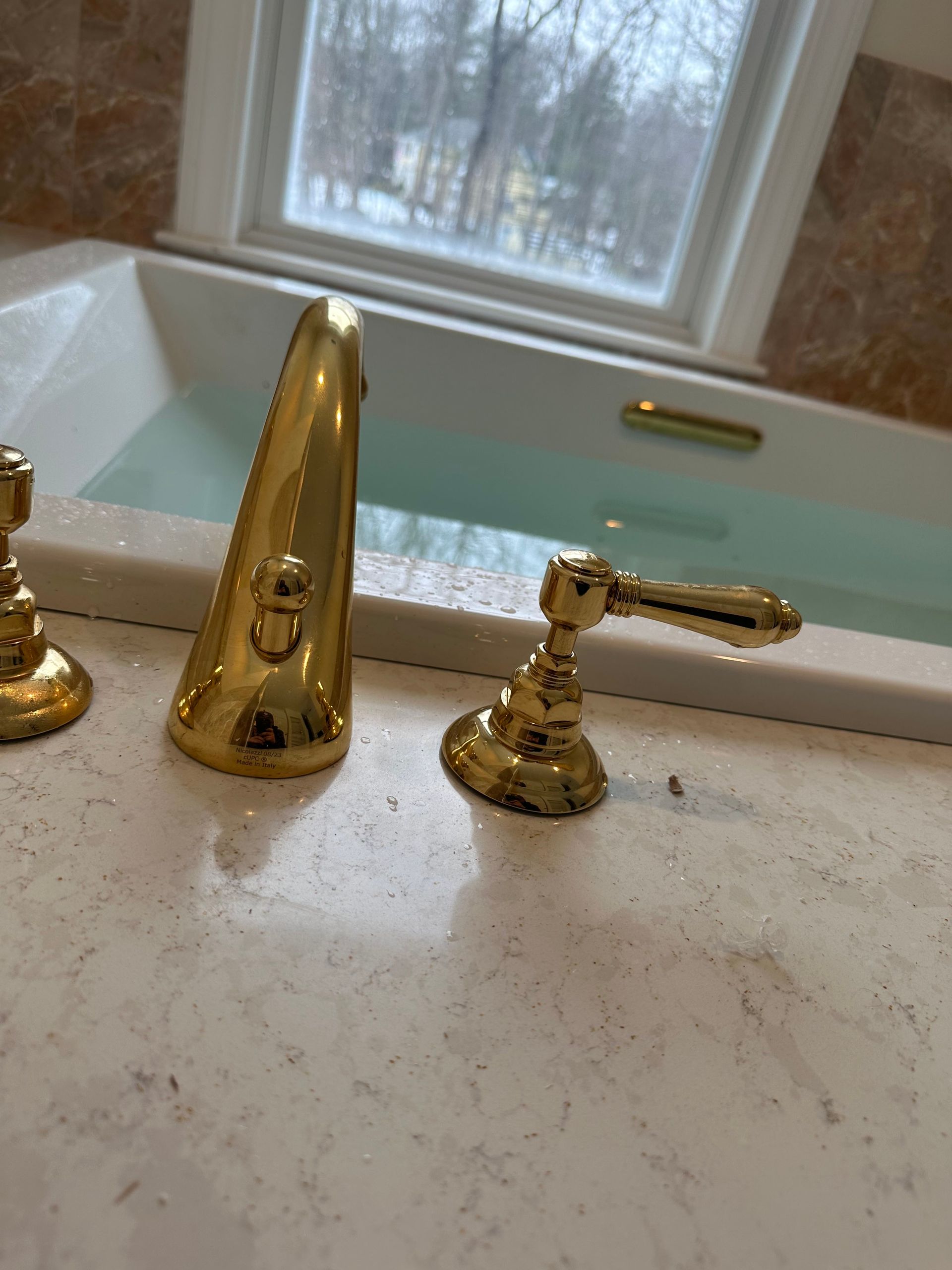 Gold faucet and handle on a bathtub with water, next to a window with snowy scenery.