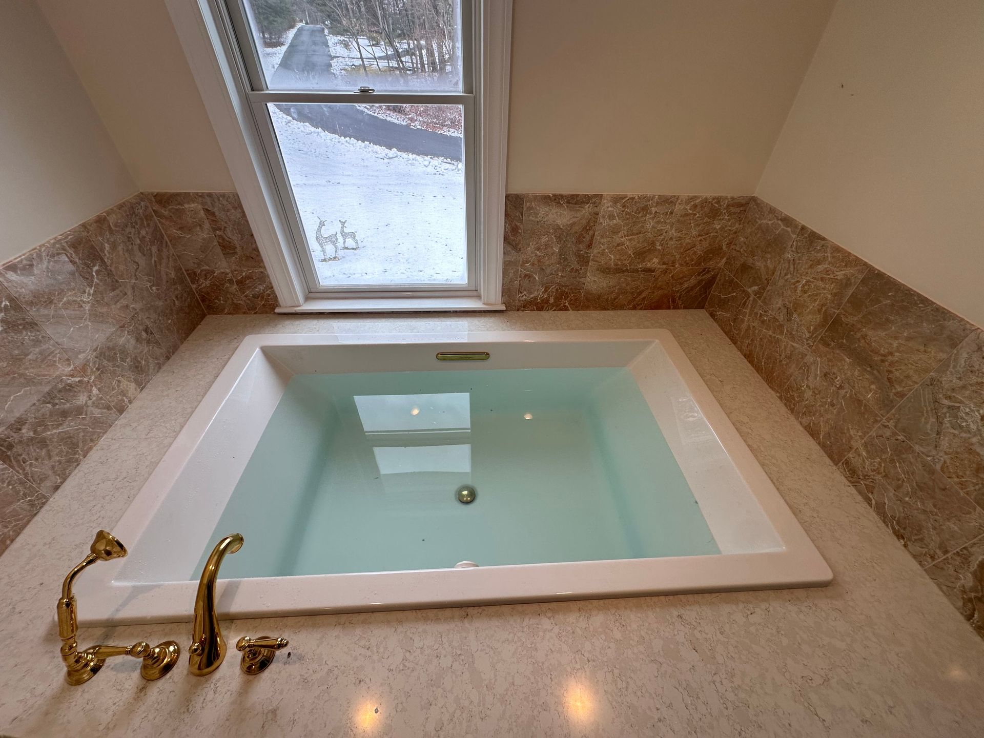 White bathtub filled with water under a window, set in beige stone, gold faucet.