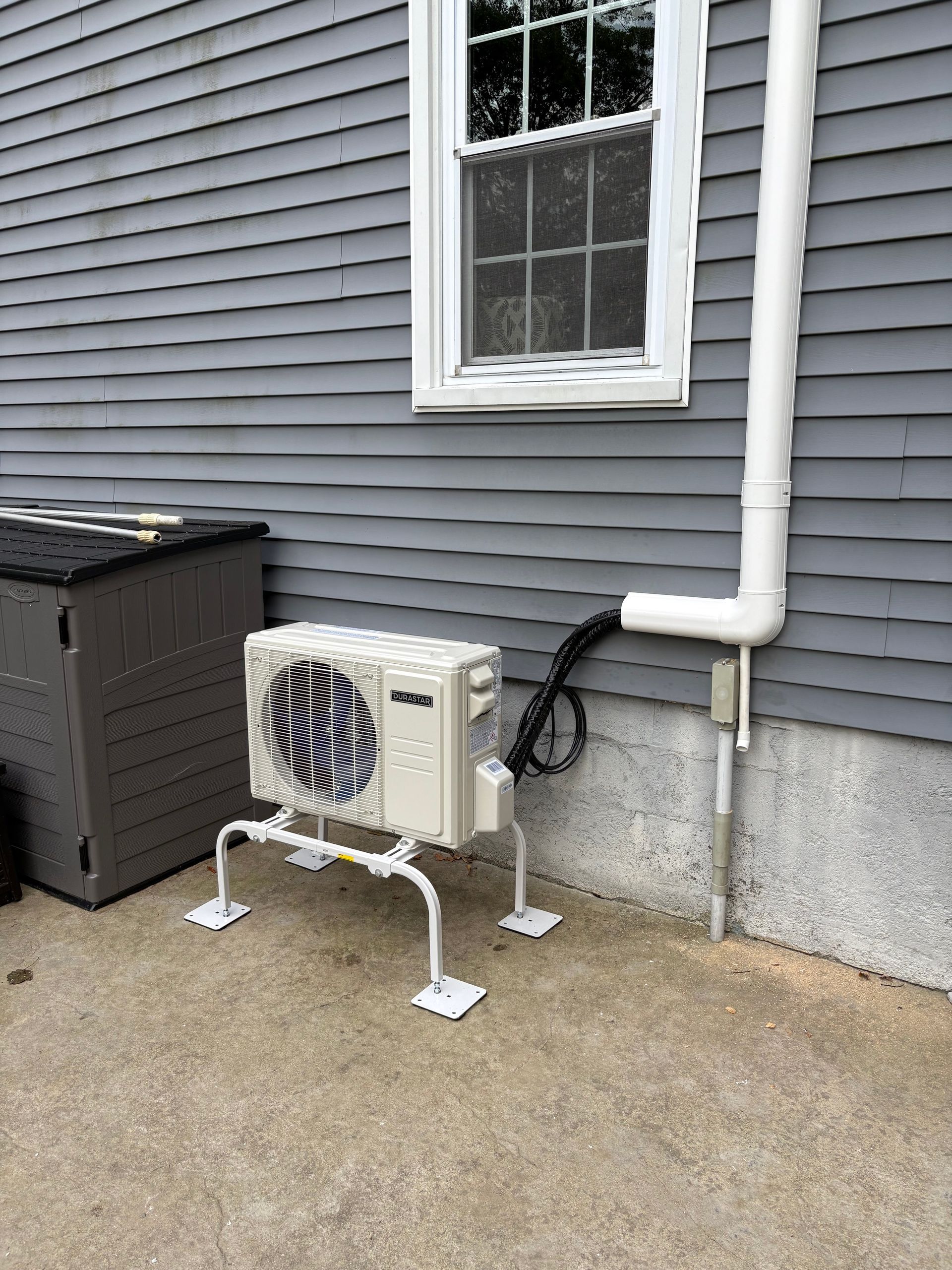 Mini-split AC unit, white, on a concrete pad outside a gray-sided building. A white drainpipe runs up the wall.