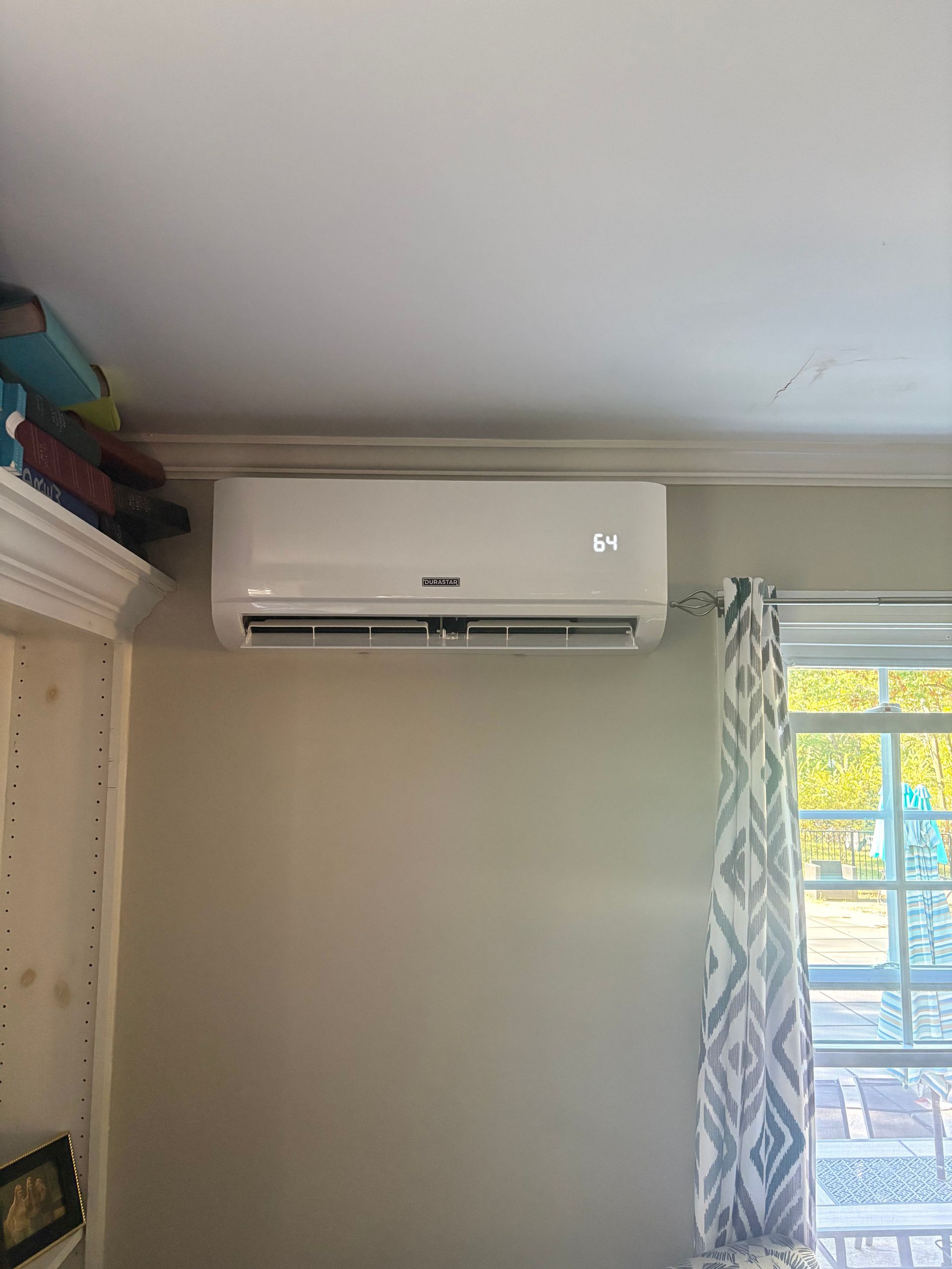 White mini-split air conditioner mounted on a beige wall, next to a window with patterned curtains.