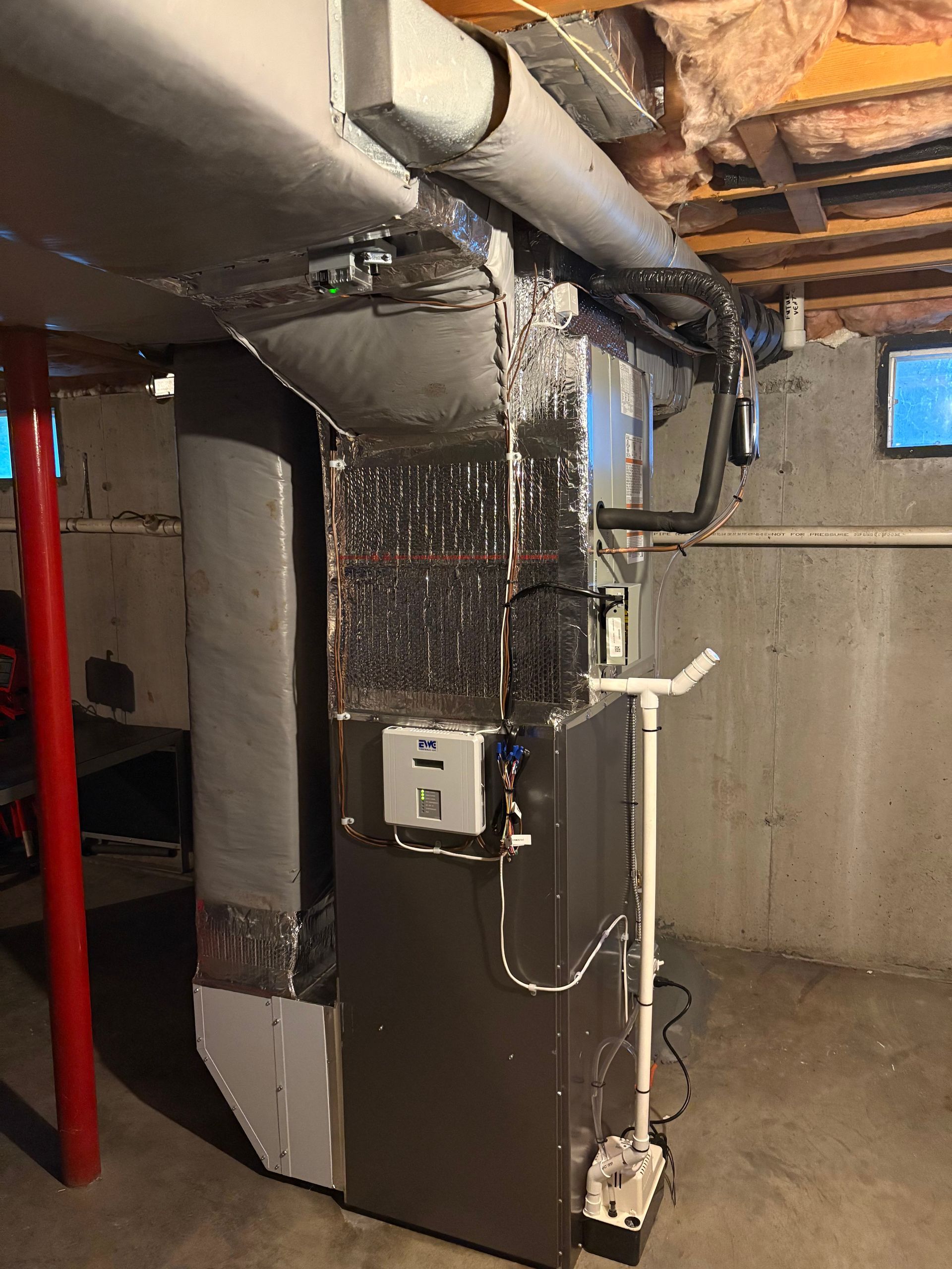 Furnace and ductwork in a basement. Grey metal unit with connected silver ducting. Concrete walls, support pole.