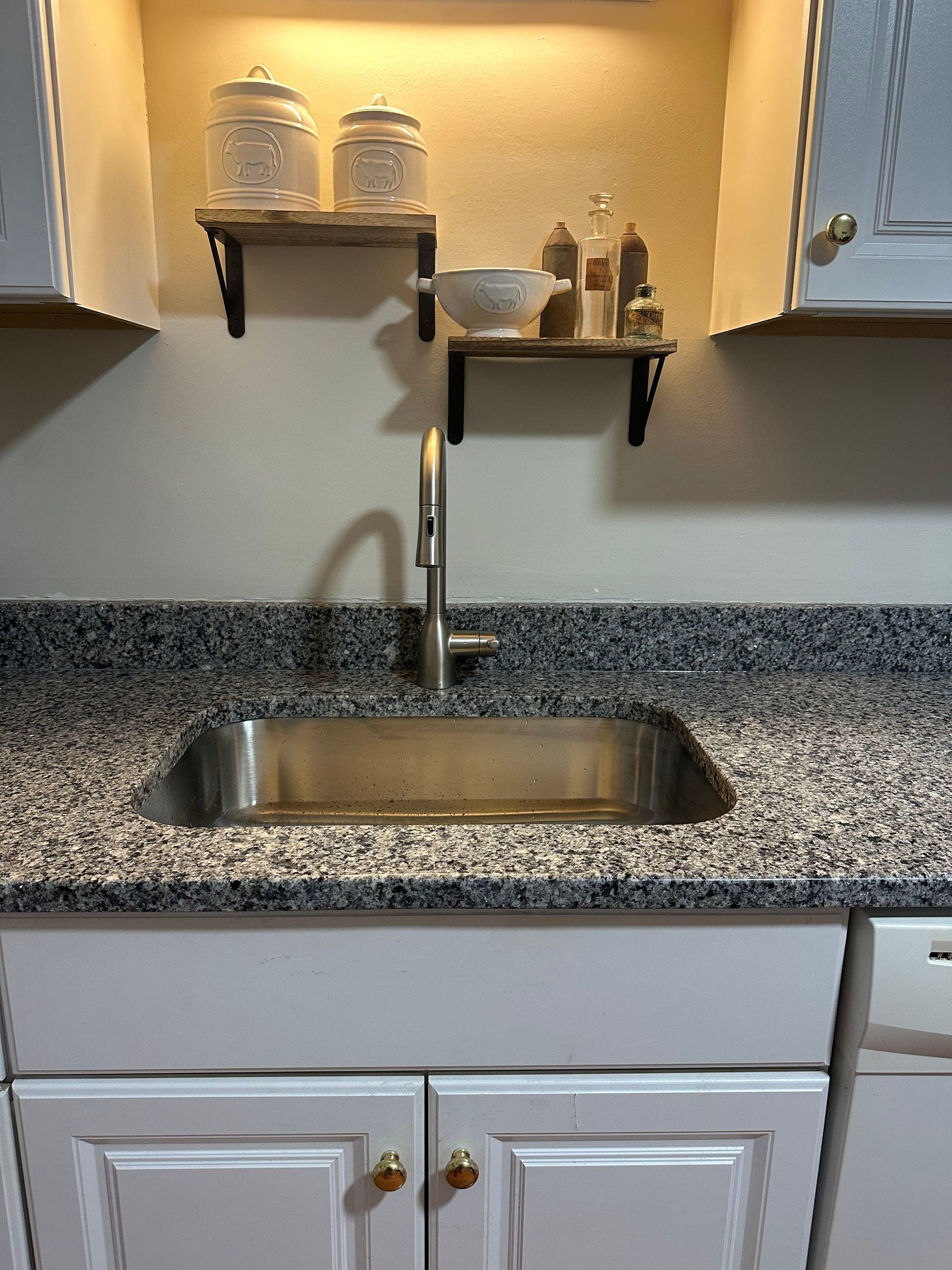 Kitchen sink with granite countertop and white cabinets, shelves with jars, and seasonings.