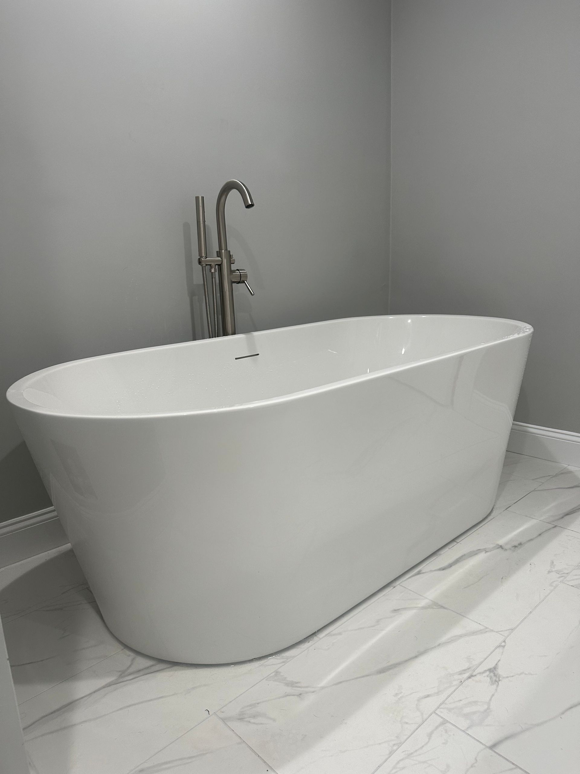White oval bathtub with tall chrome faucet against gray wall, on marble-look floor.