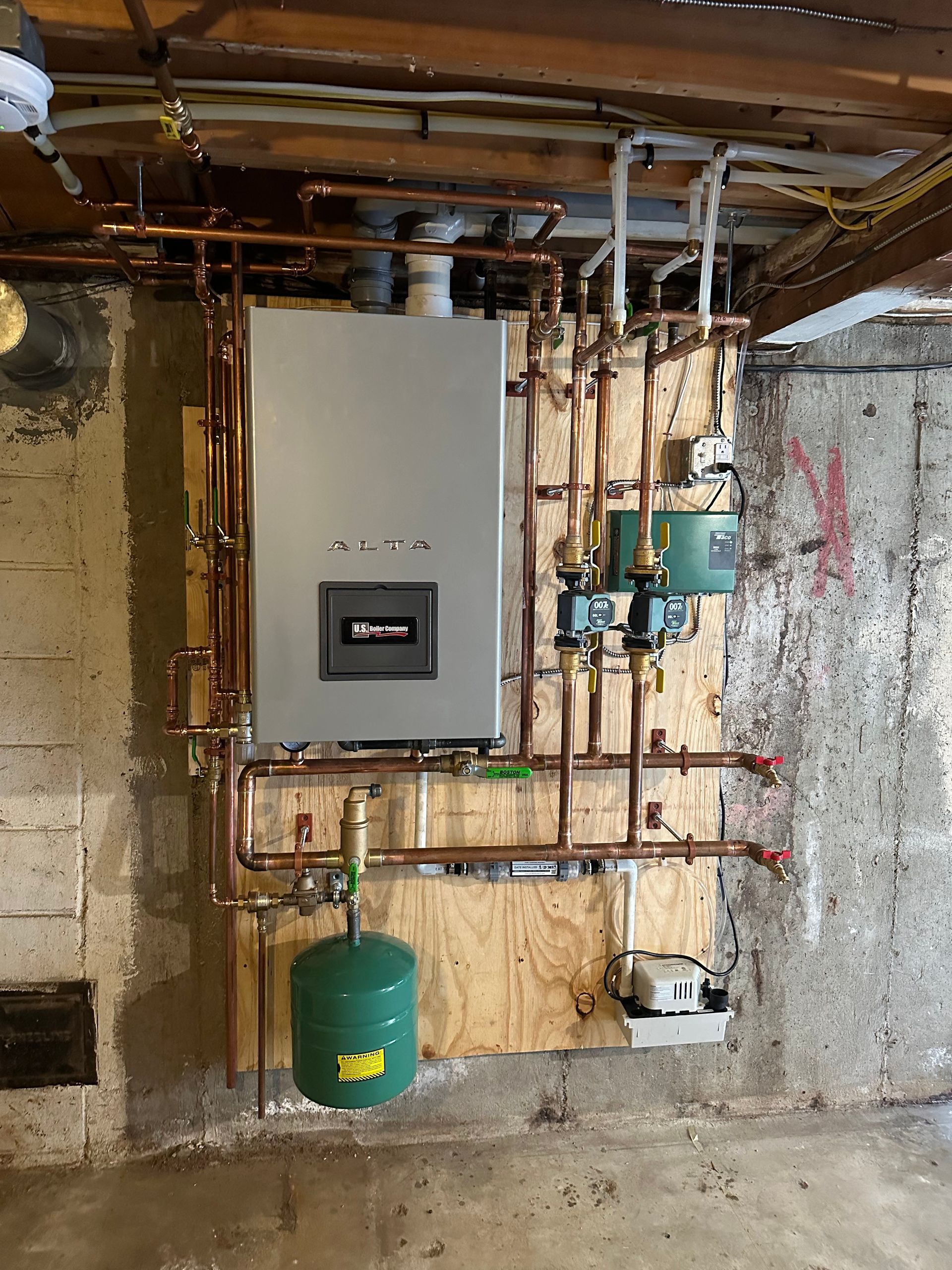A boiler system with copper pipes, a green expansion tank, and a control panel, mounted on plywood, in a basement setting.