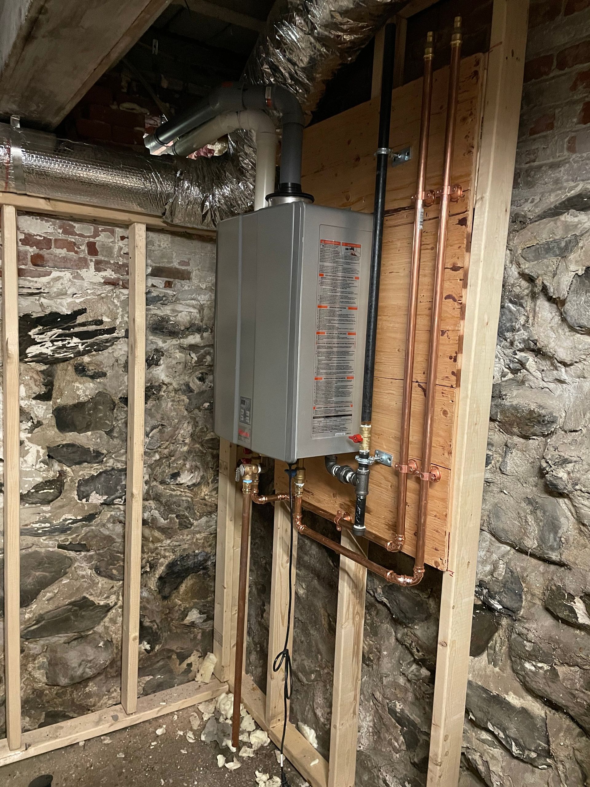 Tankless water heater installed on a wooden frame against a stone wall; copper pipes, ductwork visible.