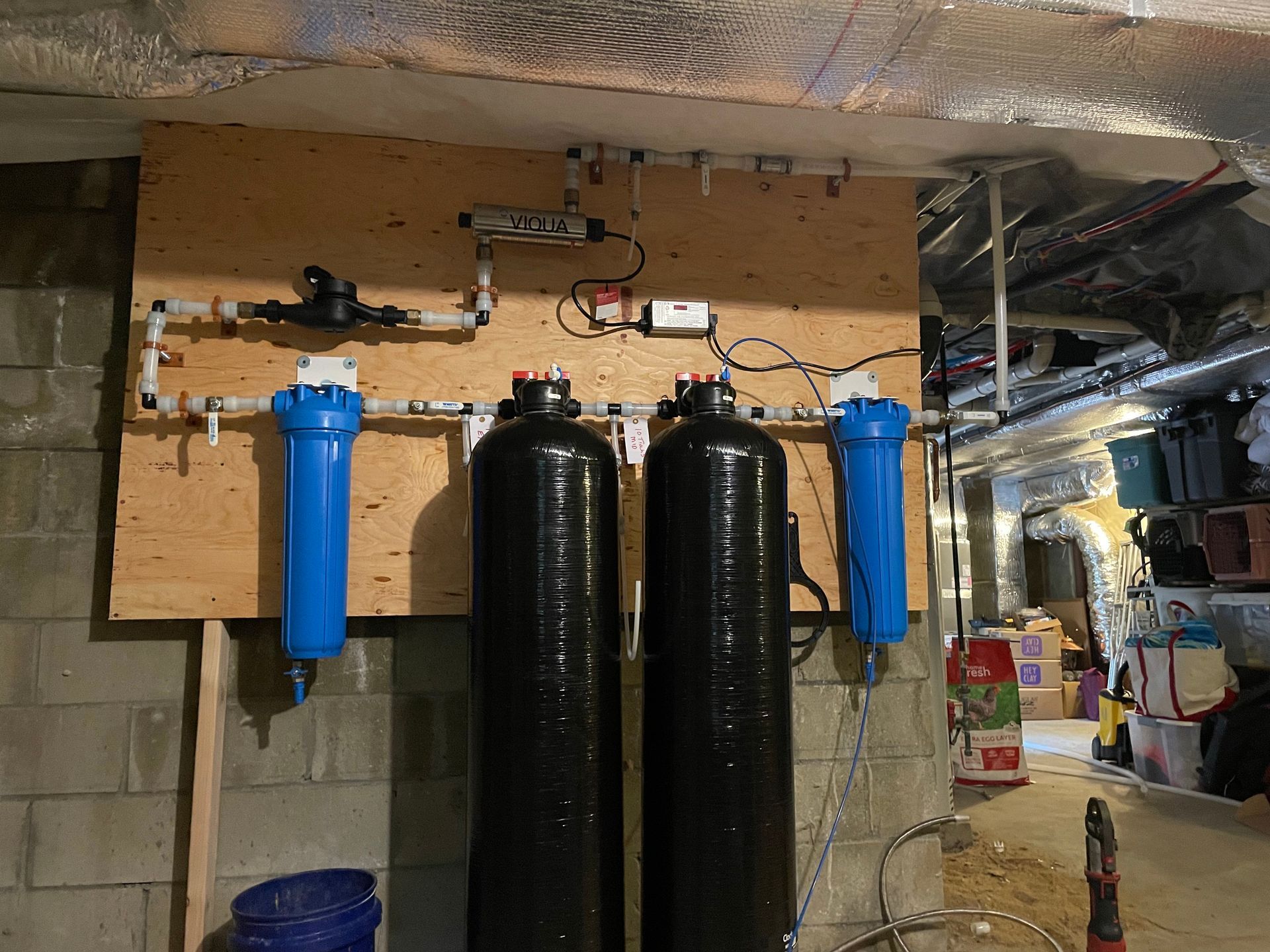 Water filtration system installed on a plywood board in a basement. Includes tanks, filters, and pipes.