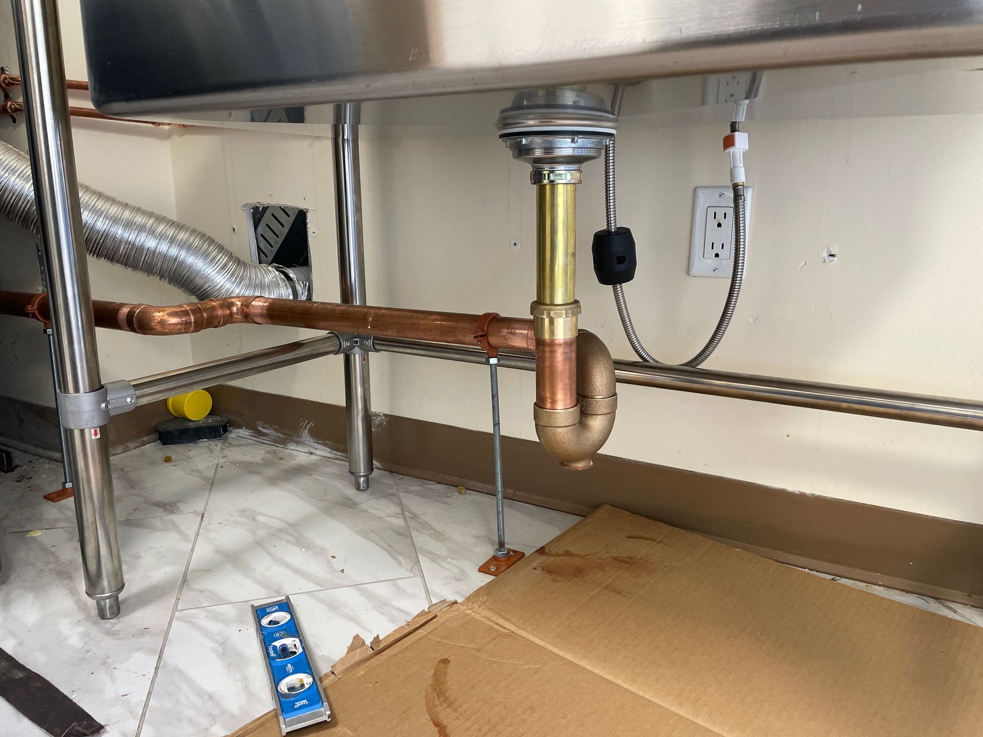 Plumbing under a stainless steel sink with copper pipes, a drain trap, and an electrical outlet on a wall.