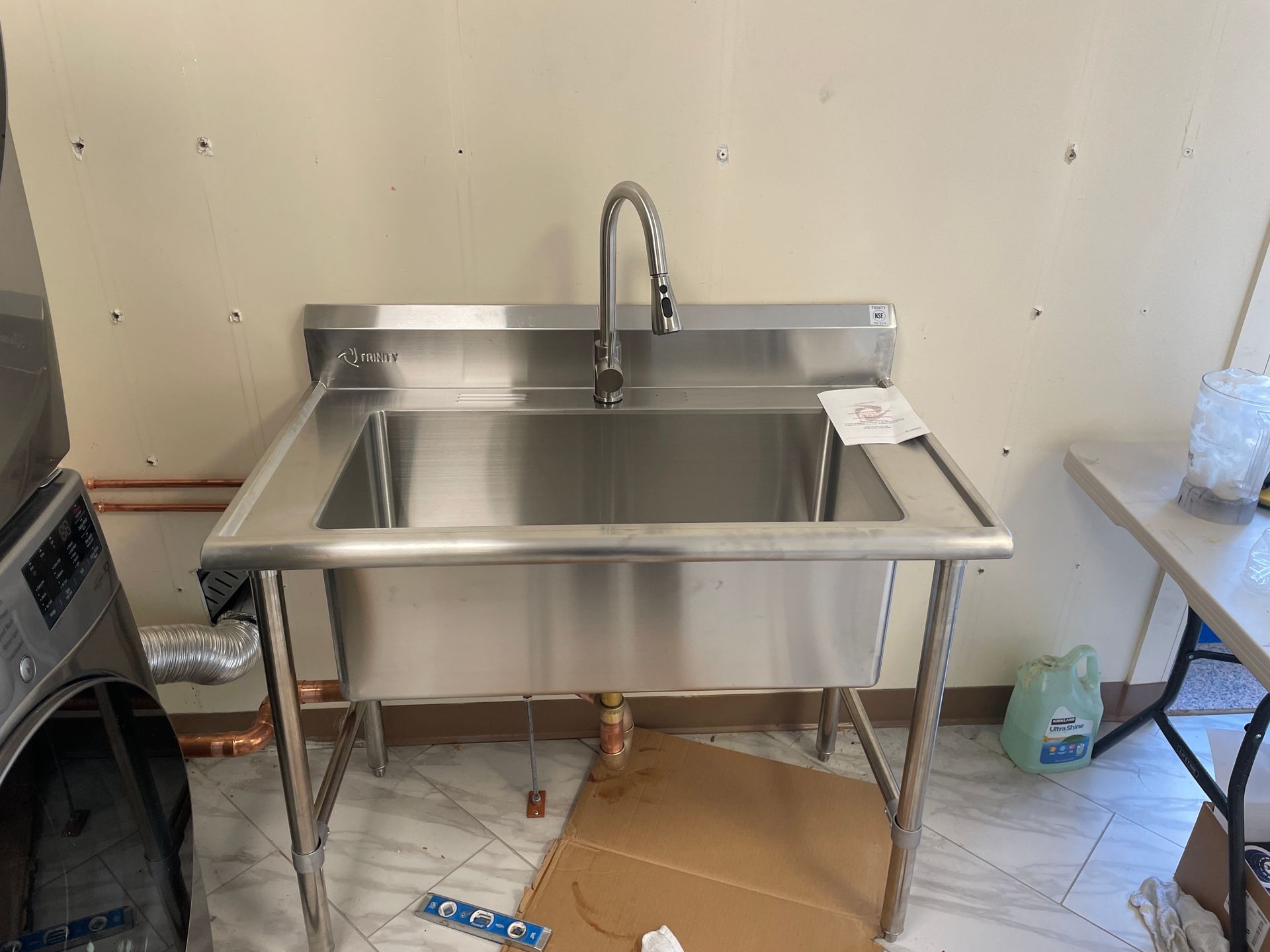 Stainless steel utility sink with a tall faucet in a room with a washing machine and table.