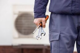 HVAC technician holding tools, standing in front of an air conditioning unit.