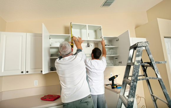 Two men are measuring cabinets in a kitchen with a tape measure.