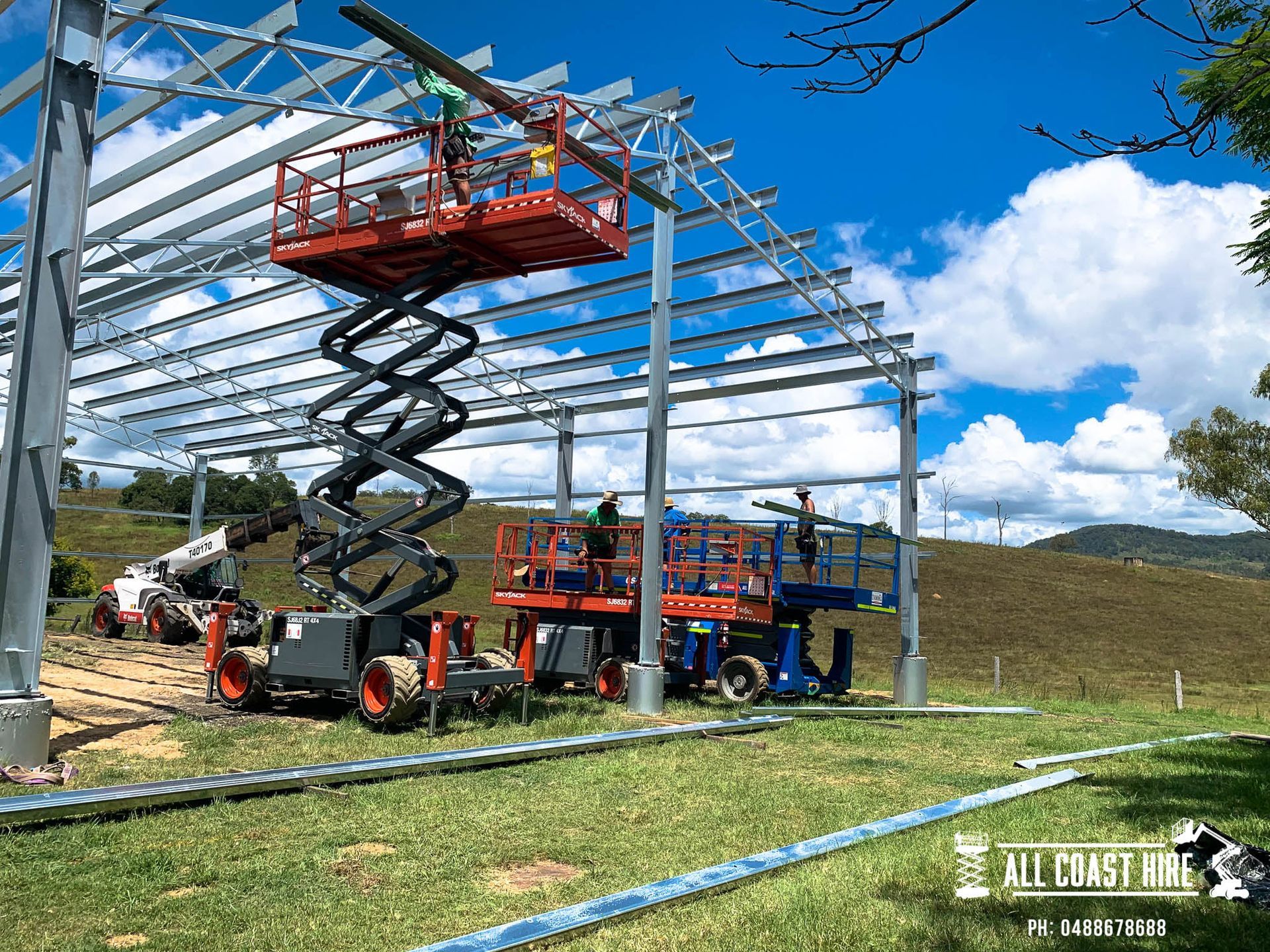 Construction workers on red and blue scissor lifts install metal roofing beams on a steel frame outdoors.