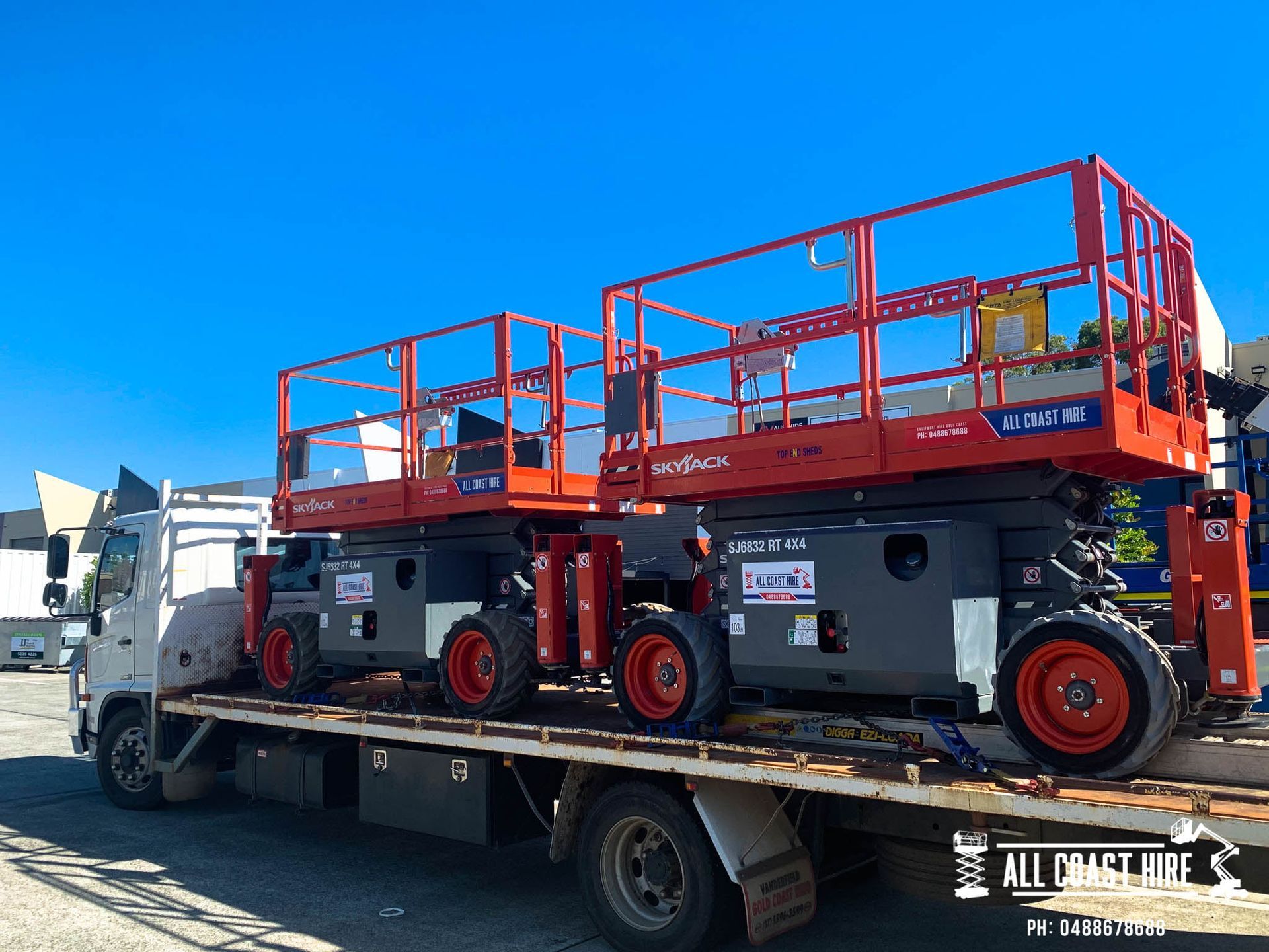 Two red and gray scissor lifts loaded on the flatbed of a white truck against a clear blue sky.