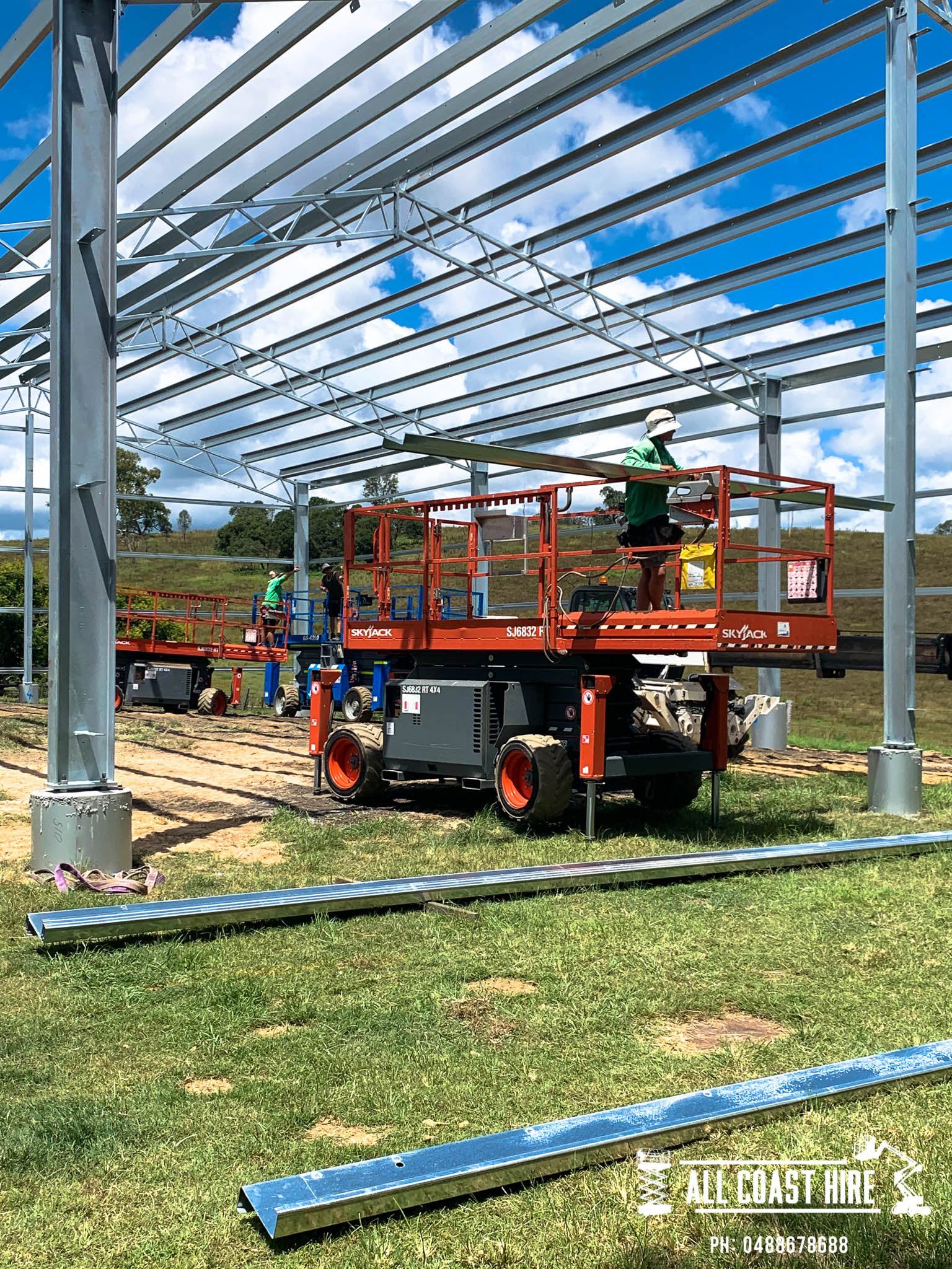 Workers on an orange scissor lift install metal roofing beams on a steel building frame in a grassy field.