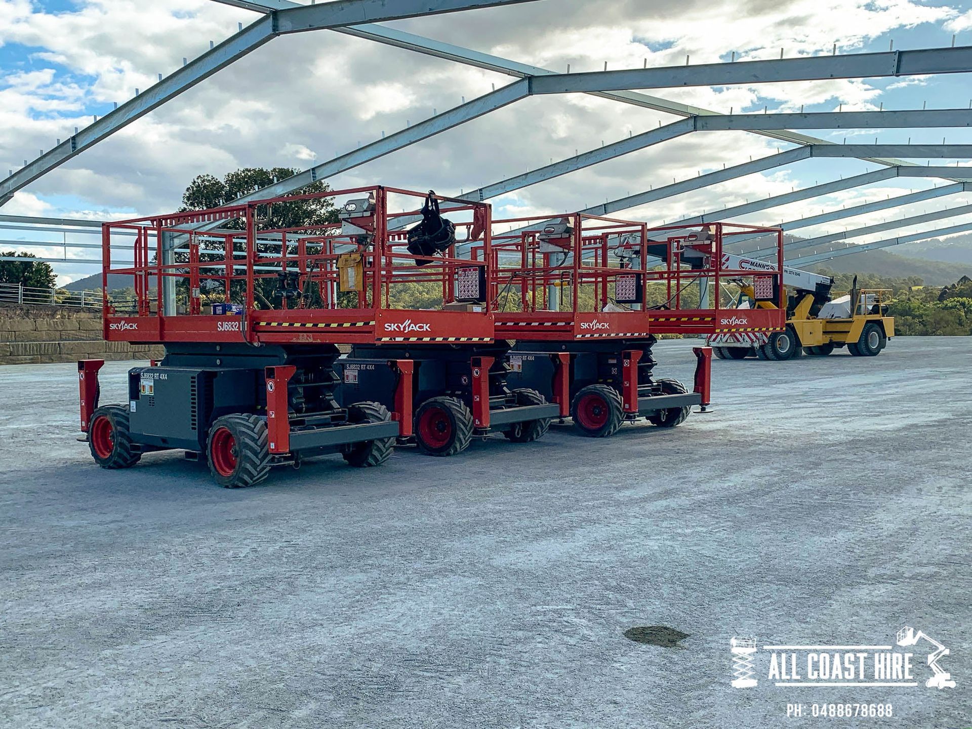 Two red scissor lifts parked on a gravel lot under a large metal building frame, with a yellow loader in the distance.