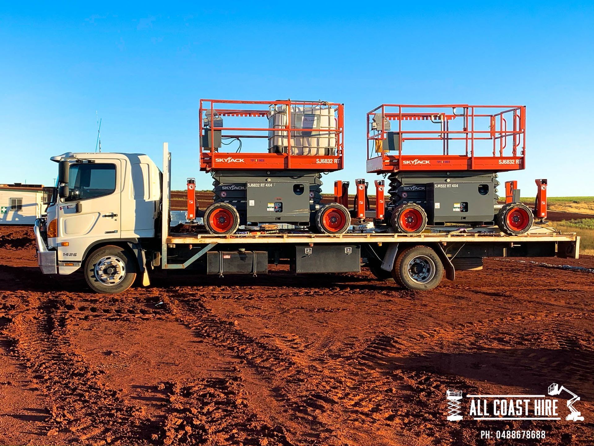 A flatbed truck transports two orange and gray scissor lifts across a red dirt construction site under a blue sky.