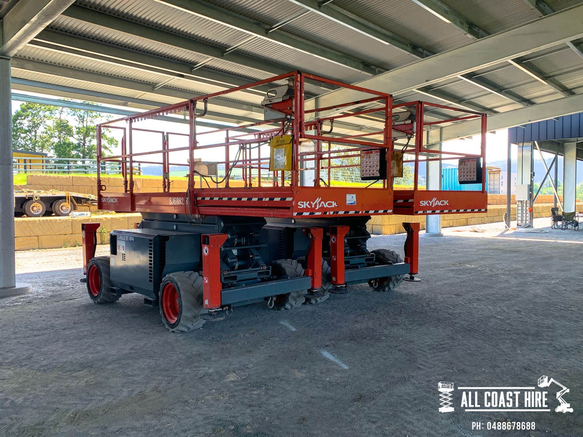 A large, red Skyjack scissor lift parked on a gravel surface under a roof structure.