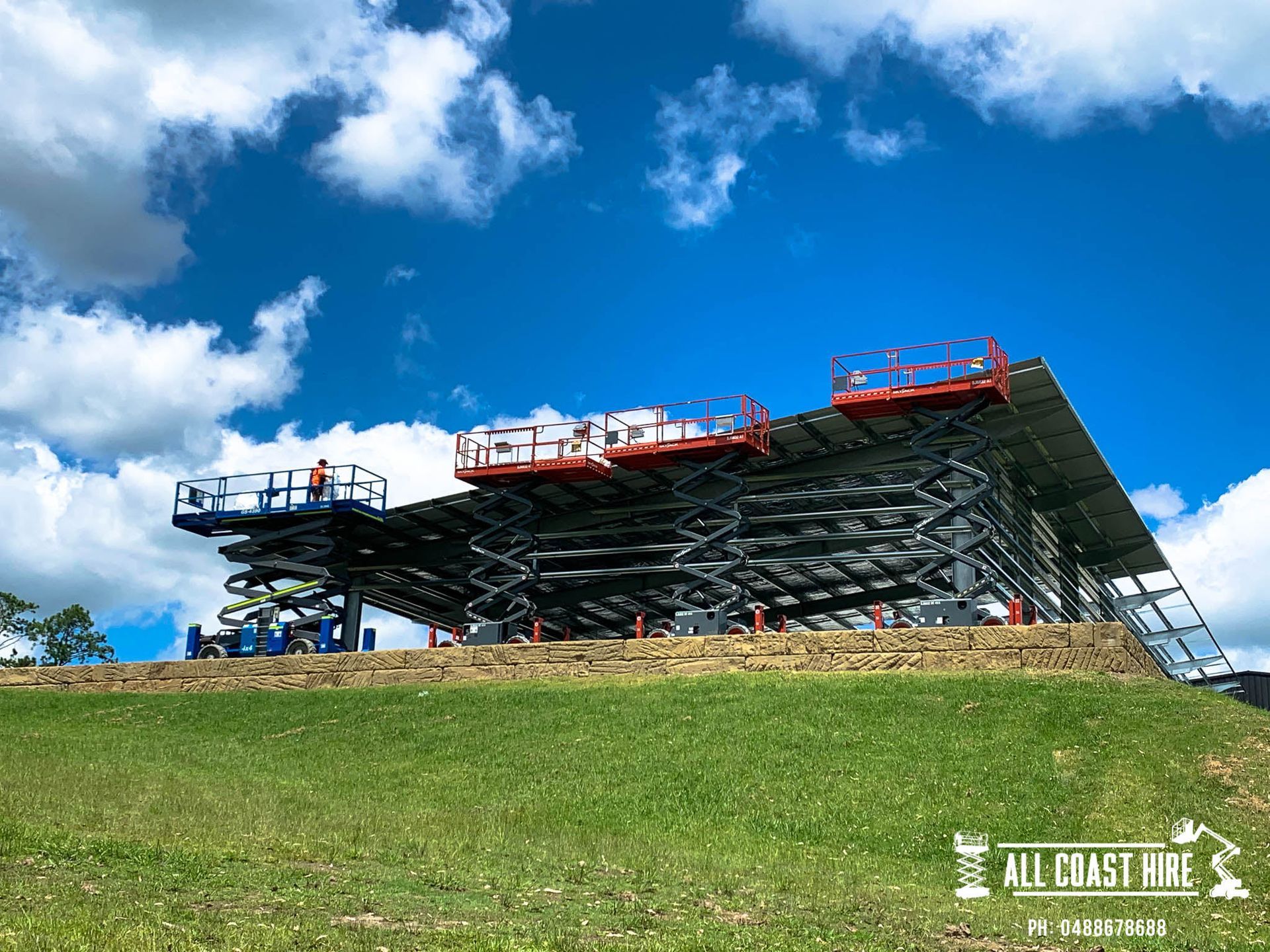 A large, industrial scissor lift structure stands on a grassy hill under a bright blue sky with white clouds.