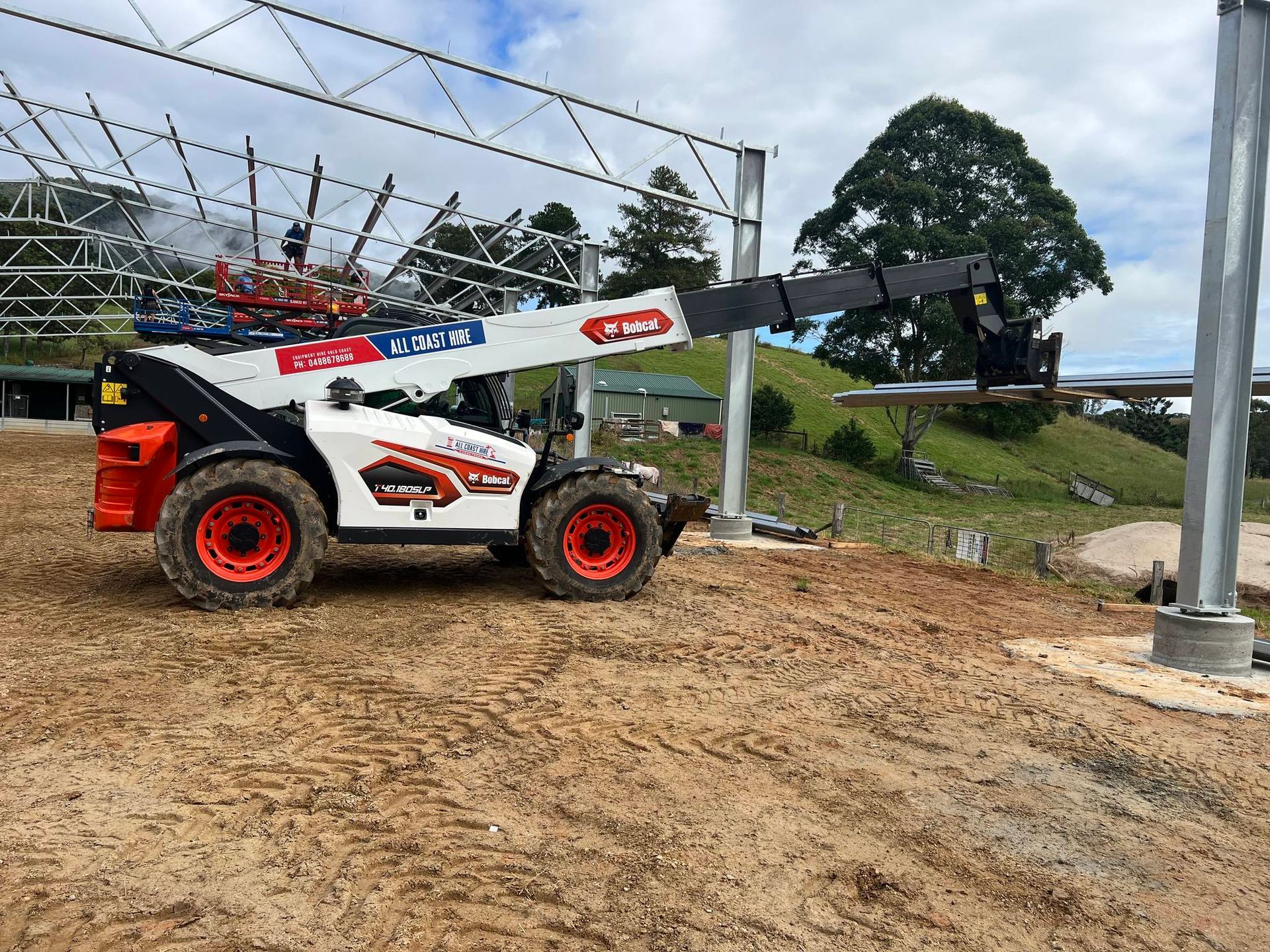 A white and orange telescopic handler lifts a structural beam at a construction site against a cloudy sky.