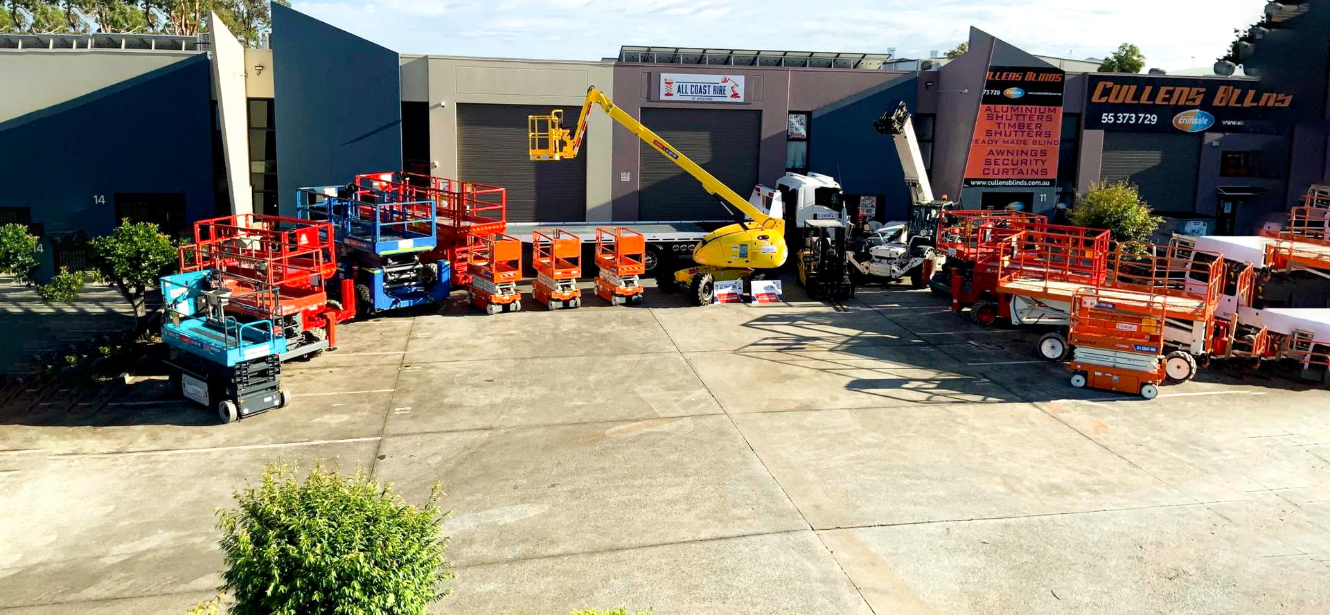 A large yard filled with various colorful construction lifts and cherry pickers parked in front of industrial buildings.
