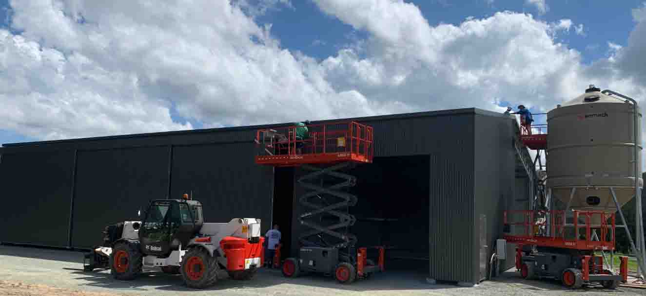 Construction workers on scissor lifts work on the exterior of a large dark-grey metal building near a feed silo.