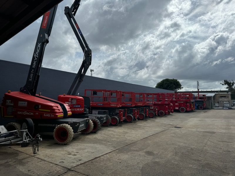 A row of red aerial work platforms and a telehandler parked on an outdoor concrete lot under a cloudy sky.