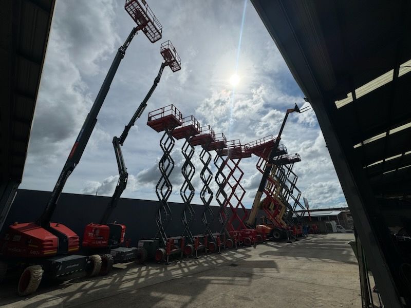 Red boom and scissor lifts parked in a row outdoors under a bright, partly cloudy sky.