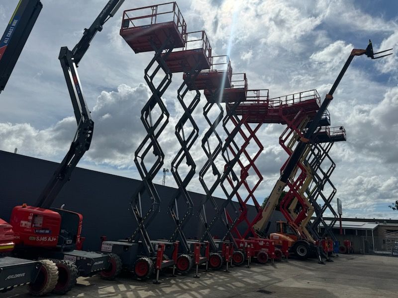A row of elevated red scissor lifts and a boom lift parked against a dark wall under a cloudy sky.
