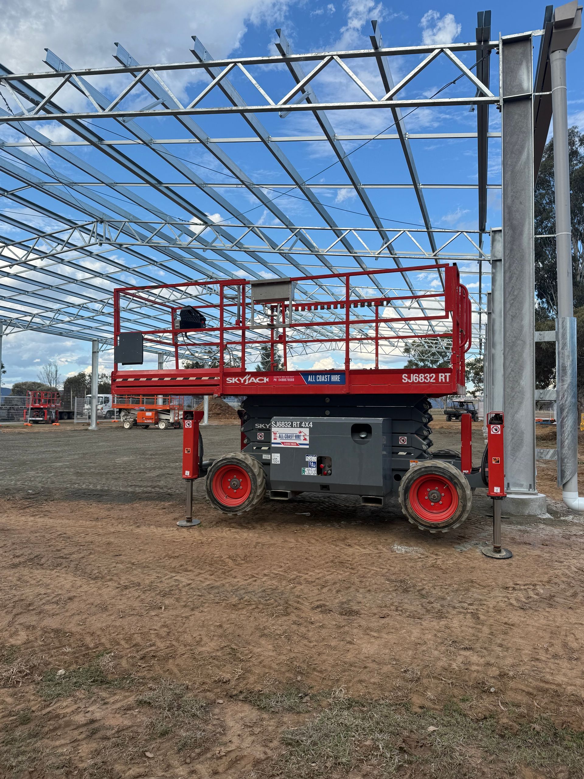 A red scissor lift parked on a dirt construction site under a large, unfinished metal roof frame.